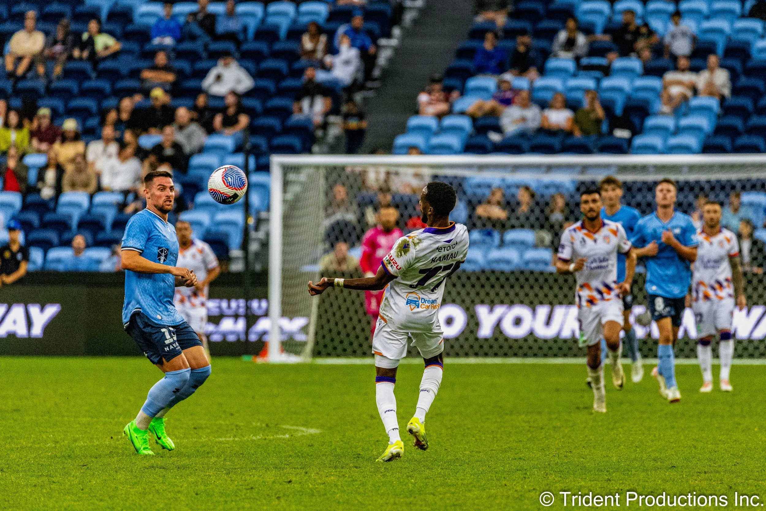 Soccer players competing for the ball on the field during a game, with spectators watching from the stands.