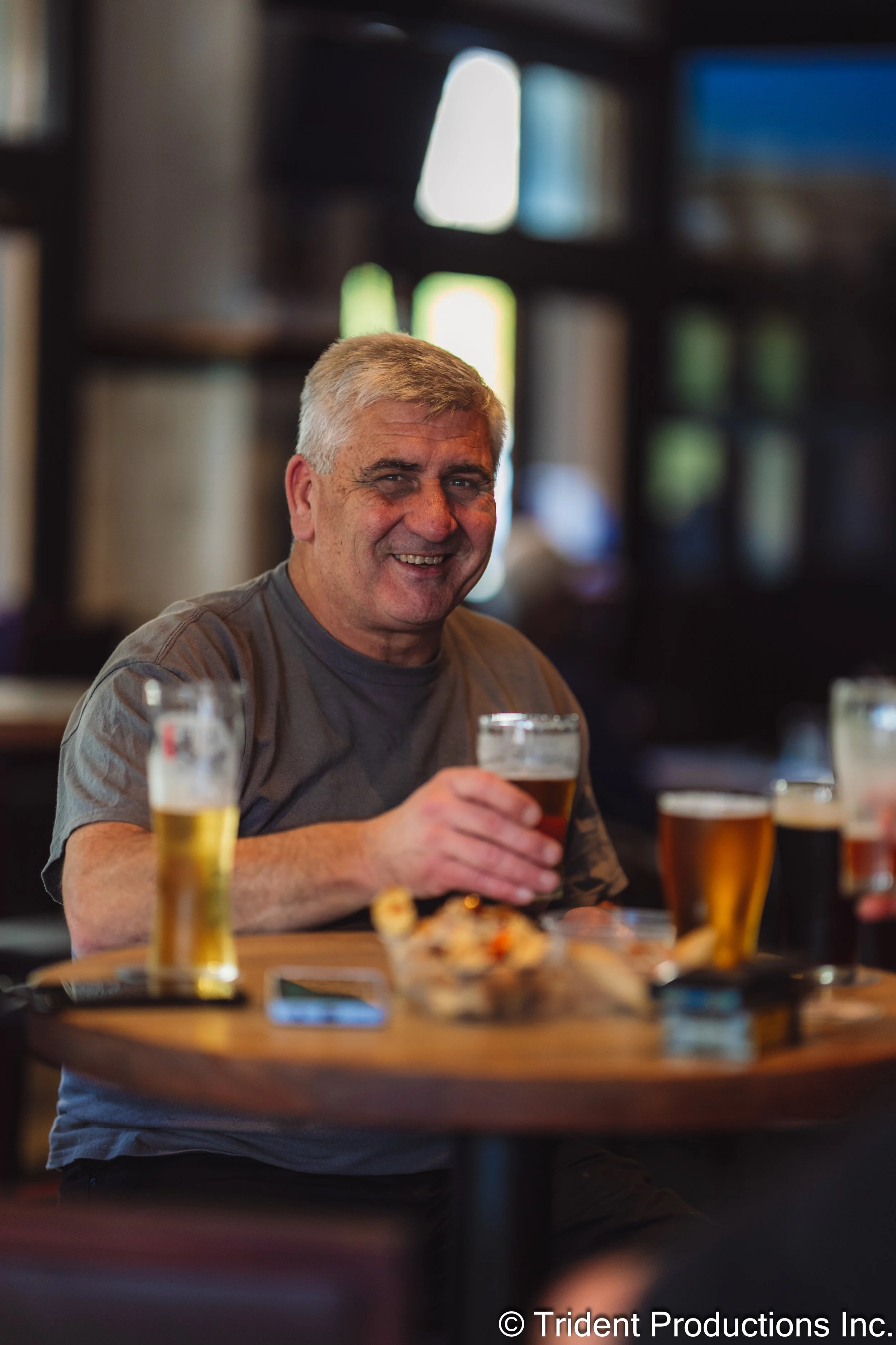 A smiling middle-aged man with gray hair sits at a wooden table, holding a glass of beer, surrounded by other glasses of beer and snacks in a cozy, dimly-lit bar or pub with blurred background.