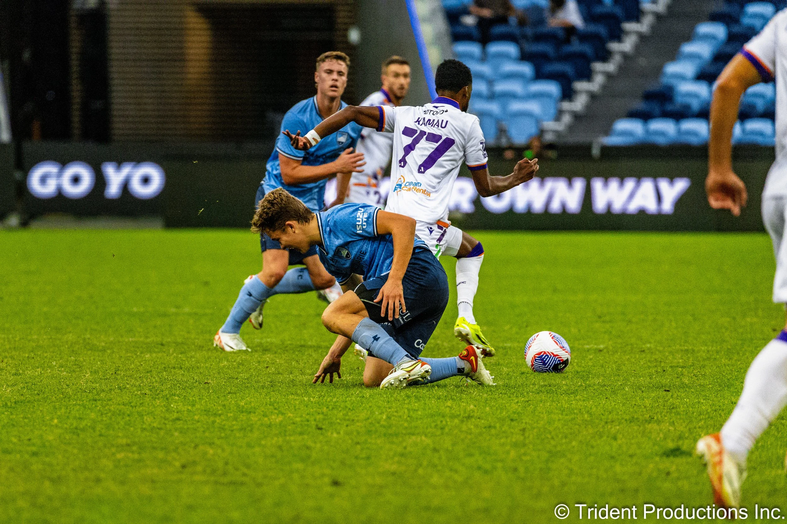 Soccer players contest for the ball during a game on a green field, with some players wearing blue uniforms and others in white uniforms with purple accents, and stadium seats in the background.