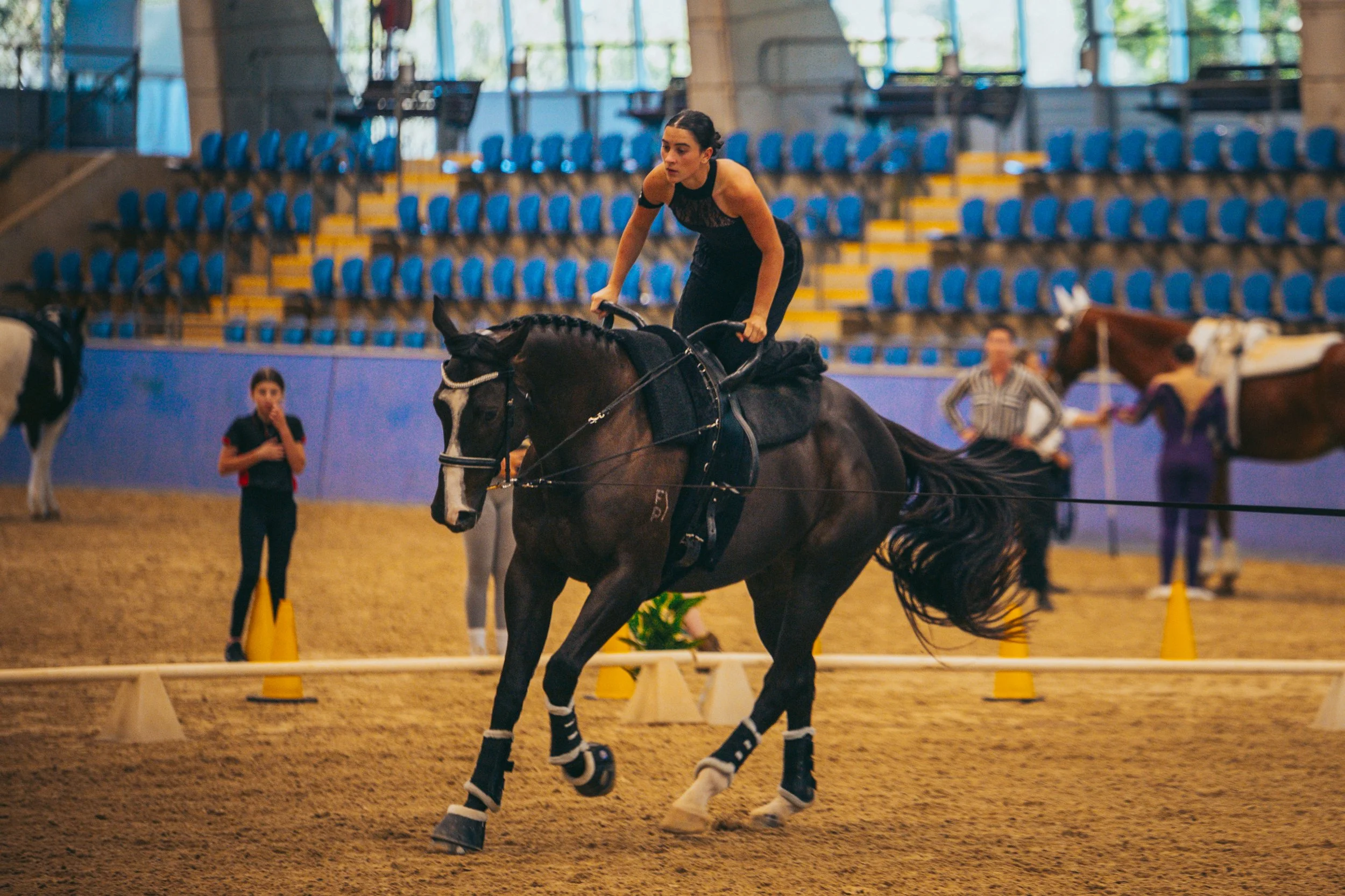 A young woman riding a horse in an indoor arena, participating in a horseback riding event, with other people and horses in the background.