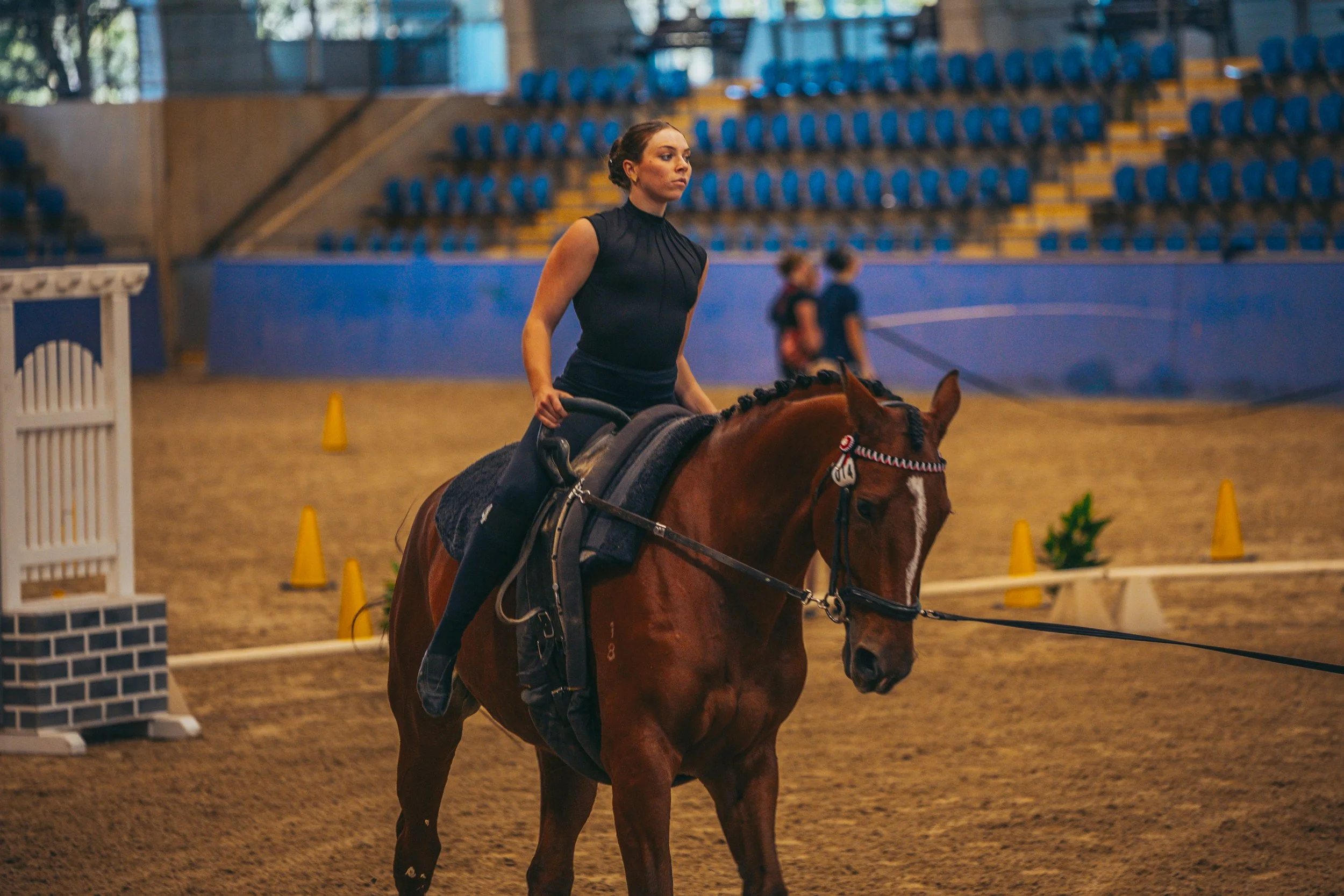 A woman riding a brown horse inside an indoor equestrian arena with yellow cones and blue seating in the background.