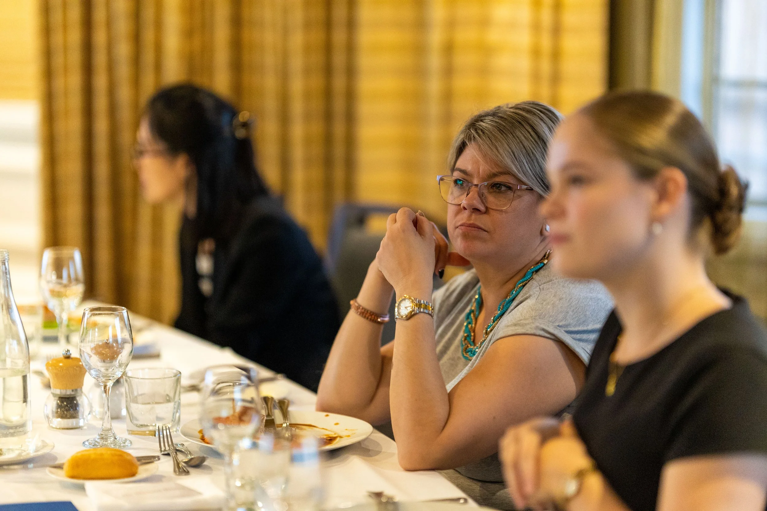 Three women sitting at a formal dining table during a meeting or event, with tableware, glasses, and a bread roll visible.