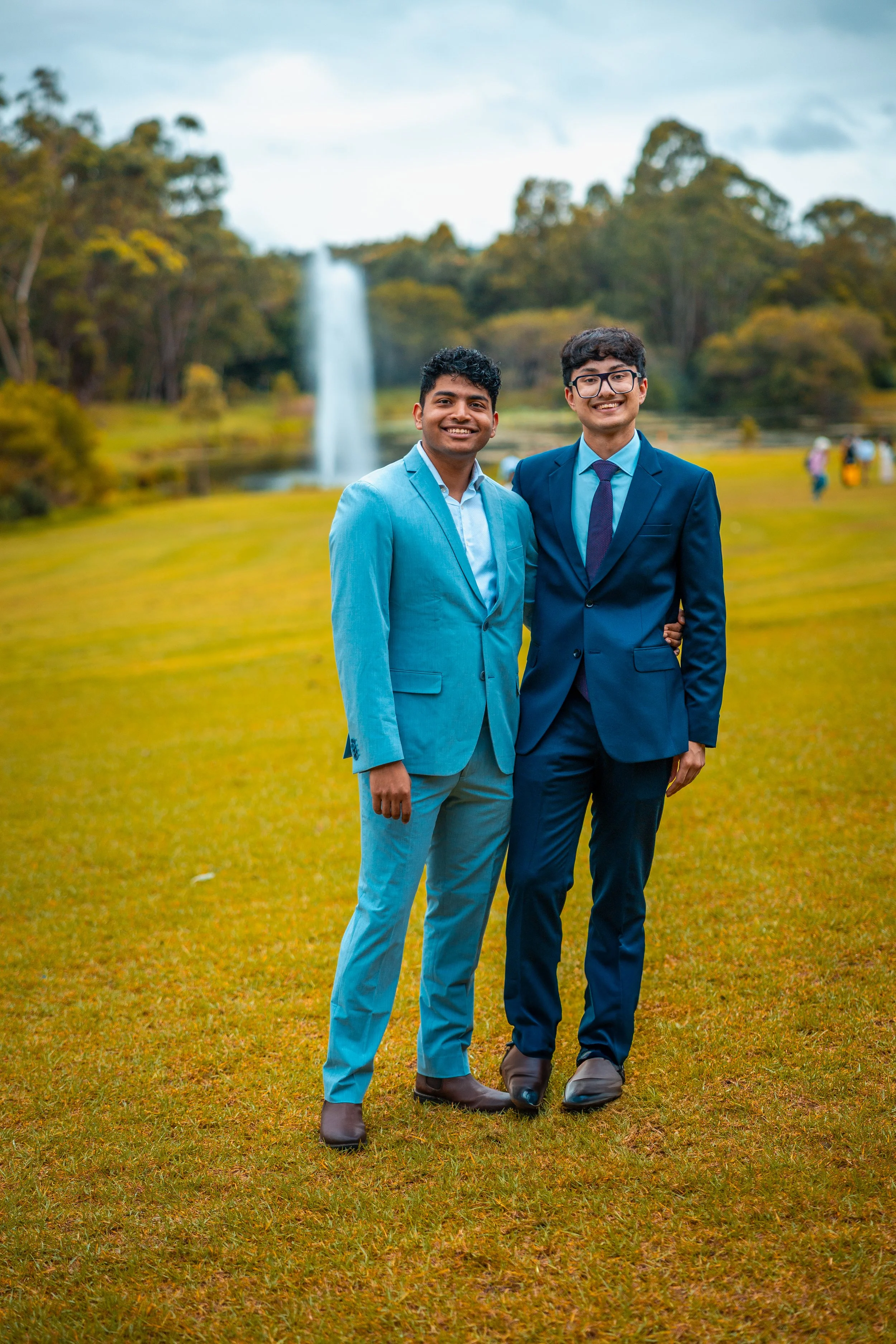 Two young men in suits standing outdoors on a grassy field with a waterfall and trees in the background.