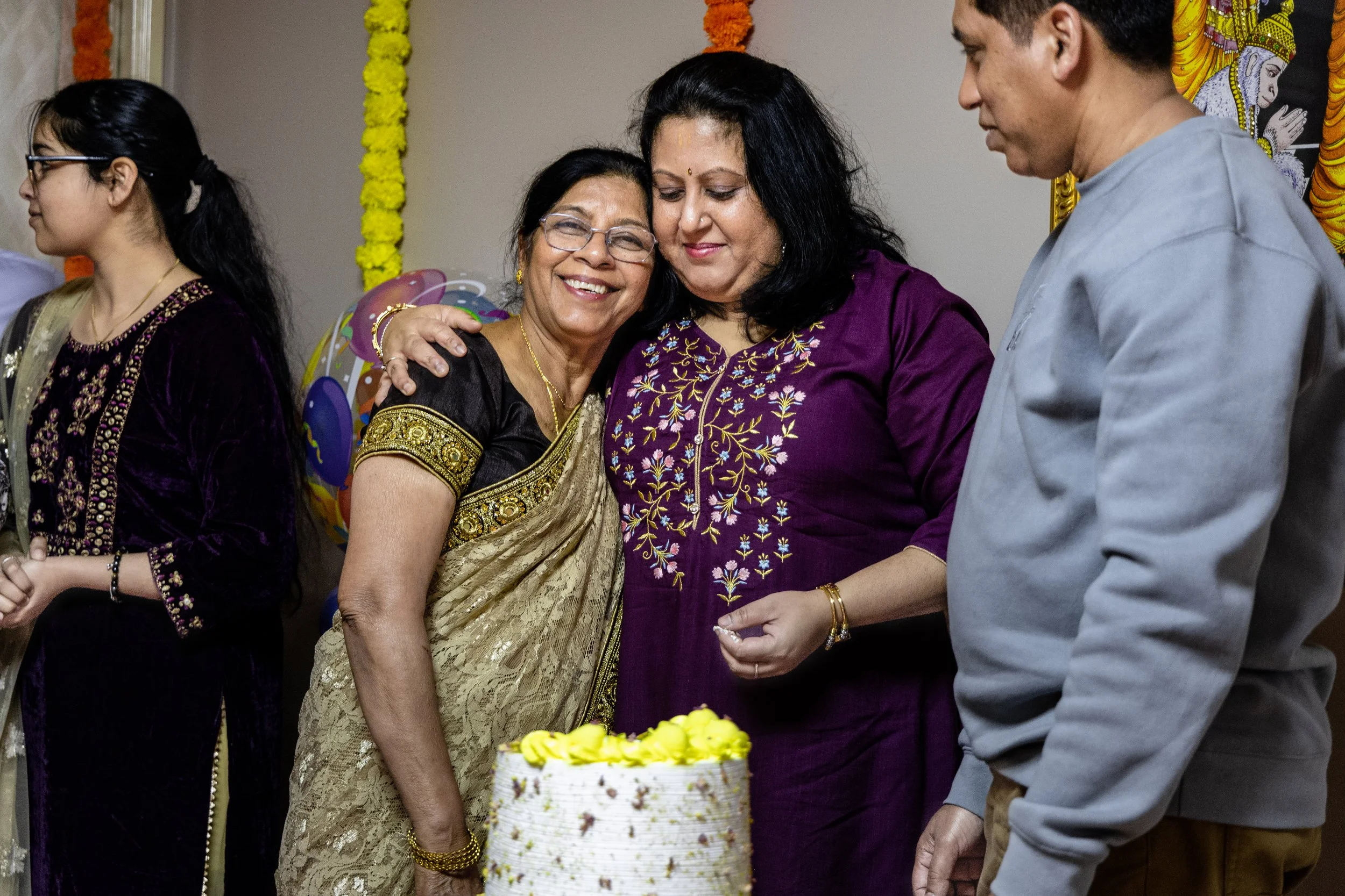 Group of people celebrating a birthday, with two women in the center sharing a hug in front of a cake, colorful decorations, and a backdrop featuring a Hindu deity.