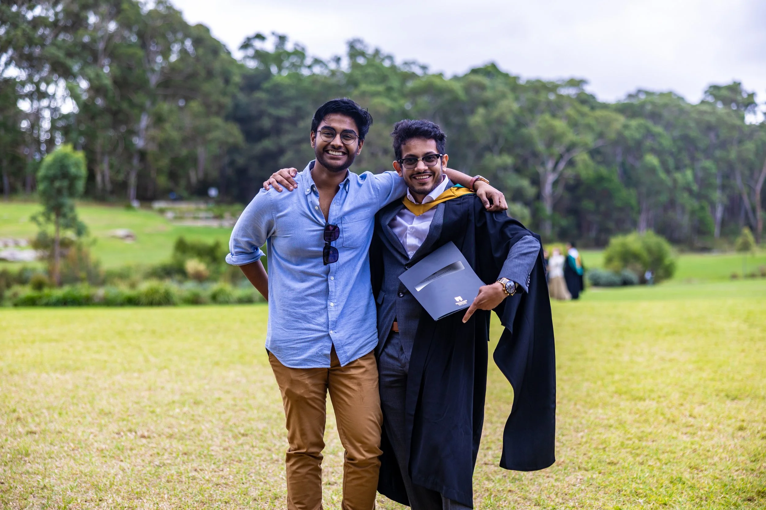 Two young men standing outdoors on a grassy field, one in graduation gown and cap holding a diploma, the other in casual clothes with sunglasses hanging from his shirt, smiling and hugging.
