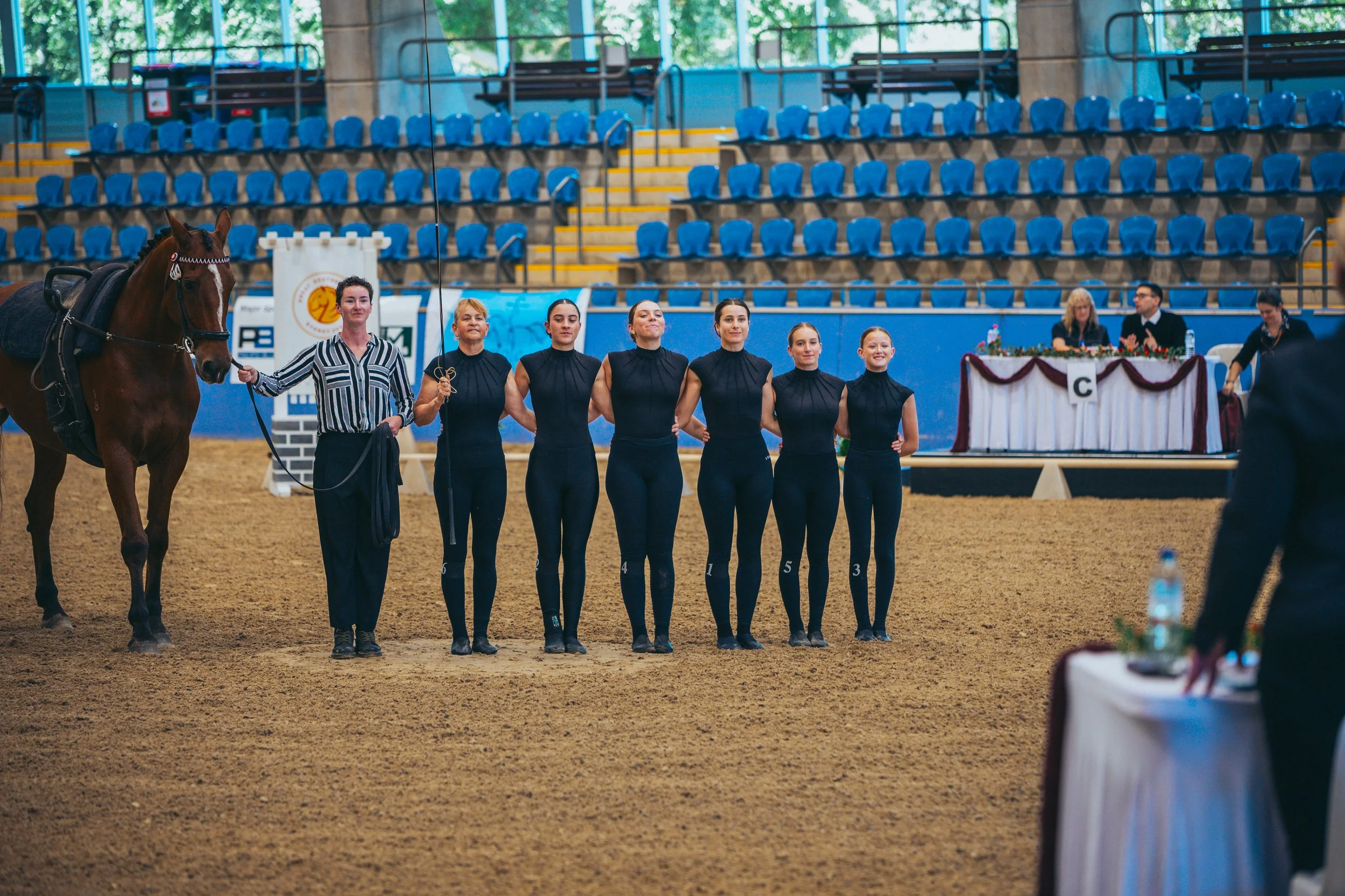 Six women and one woman holding a horse inside an indoor arena with blue seating in the background, during a competition or event.