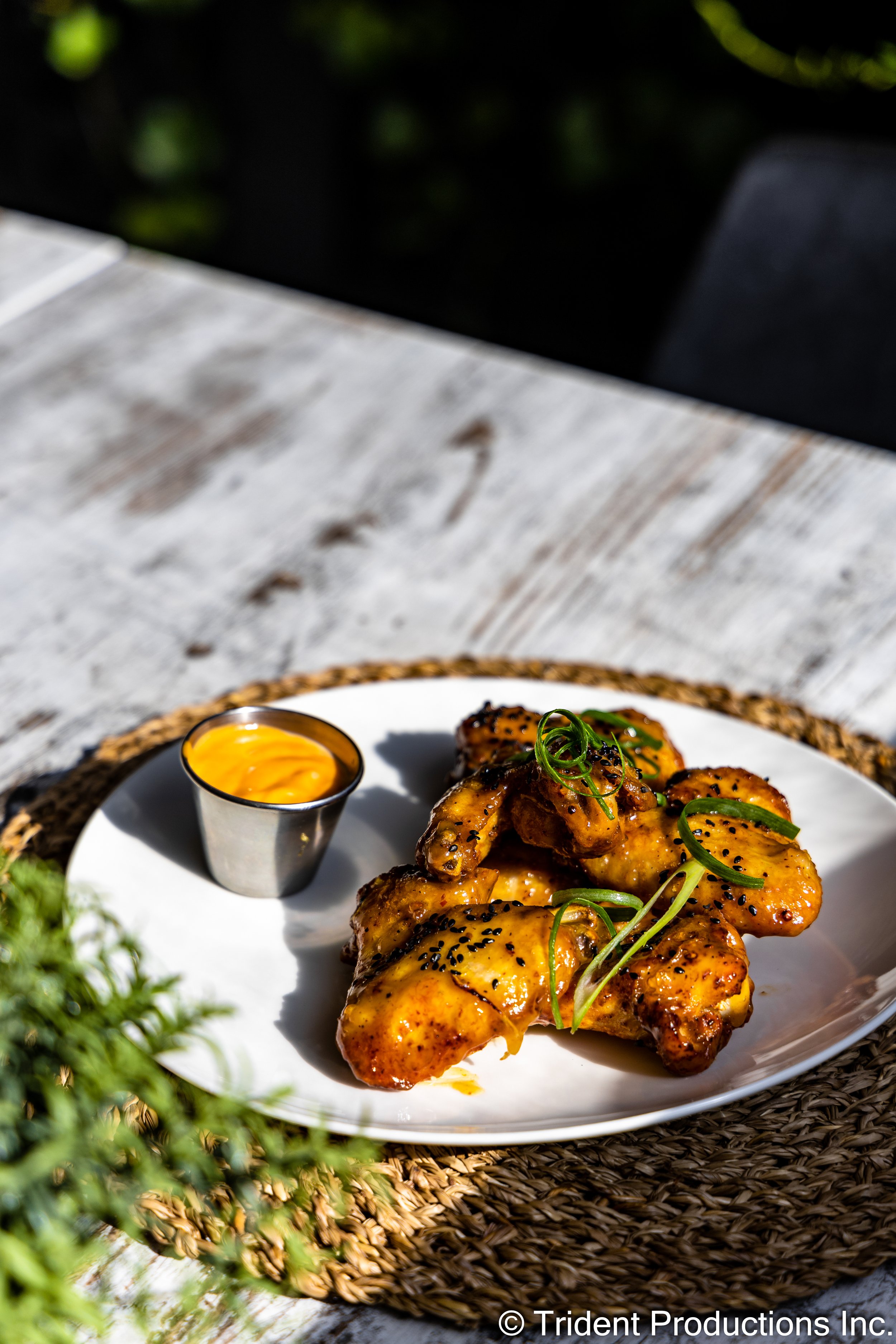 Plate of cooked chicken wings garnished with green onion, served with a side of dipping sauce in a small metal container on a woven placemat outdoors.