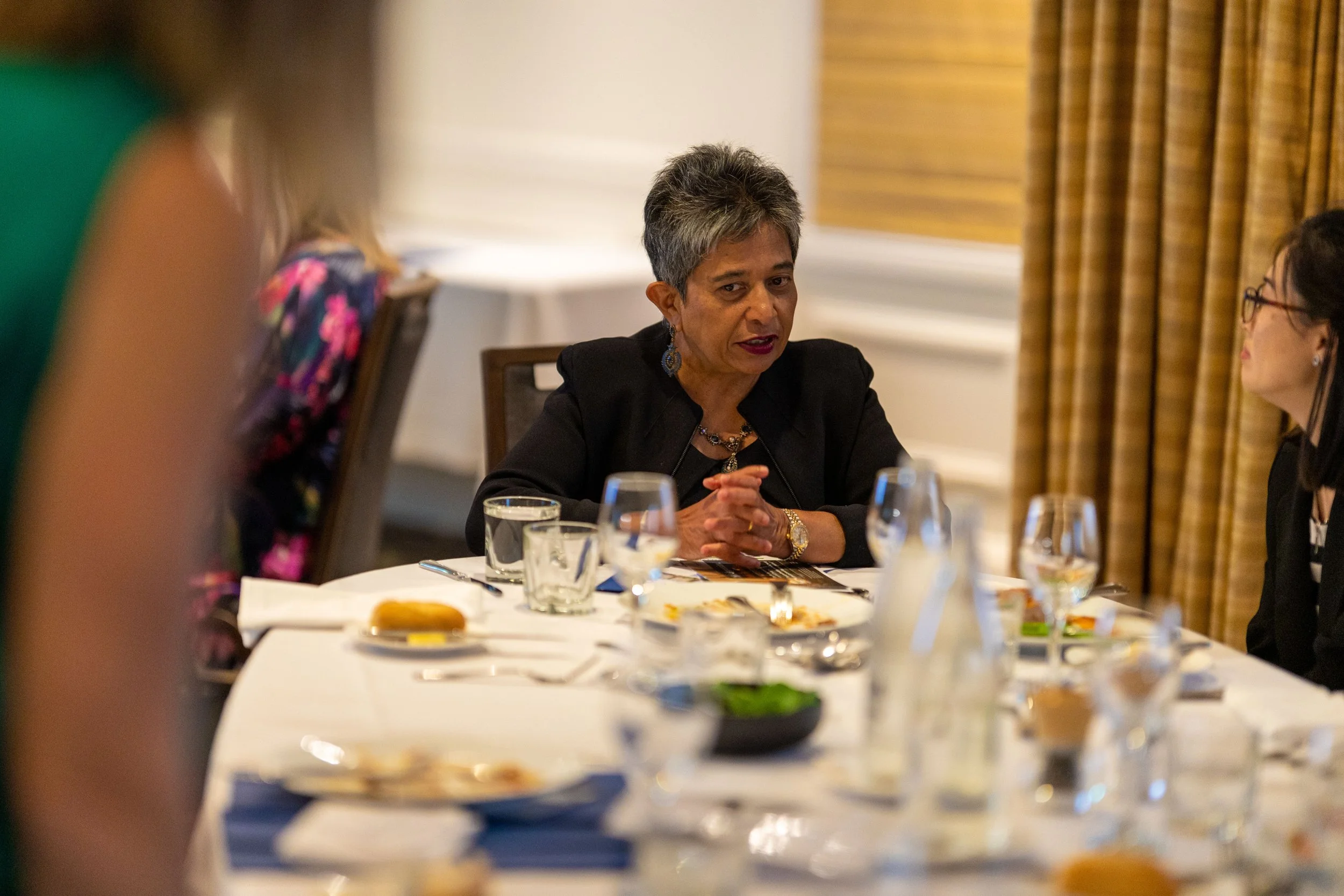 A woman with short gray hair wearing a black blazer and earrings is seated at a dining table, engaging in conversation with a woman on her right, with plates, glasses, and food on the table in a warmly lit room with beige walls and curtains.