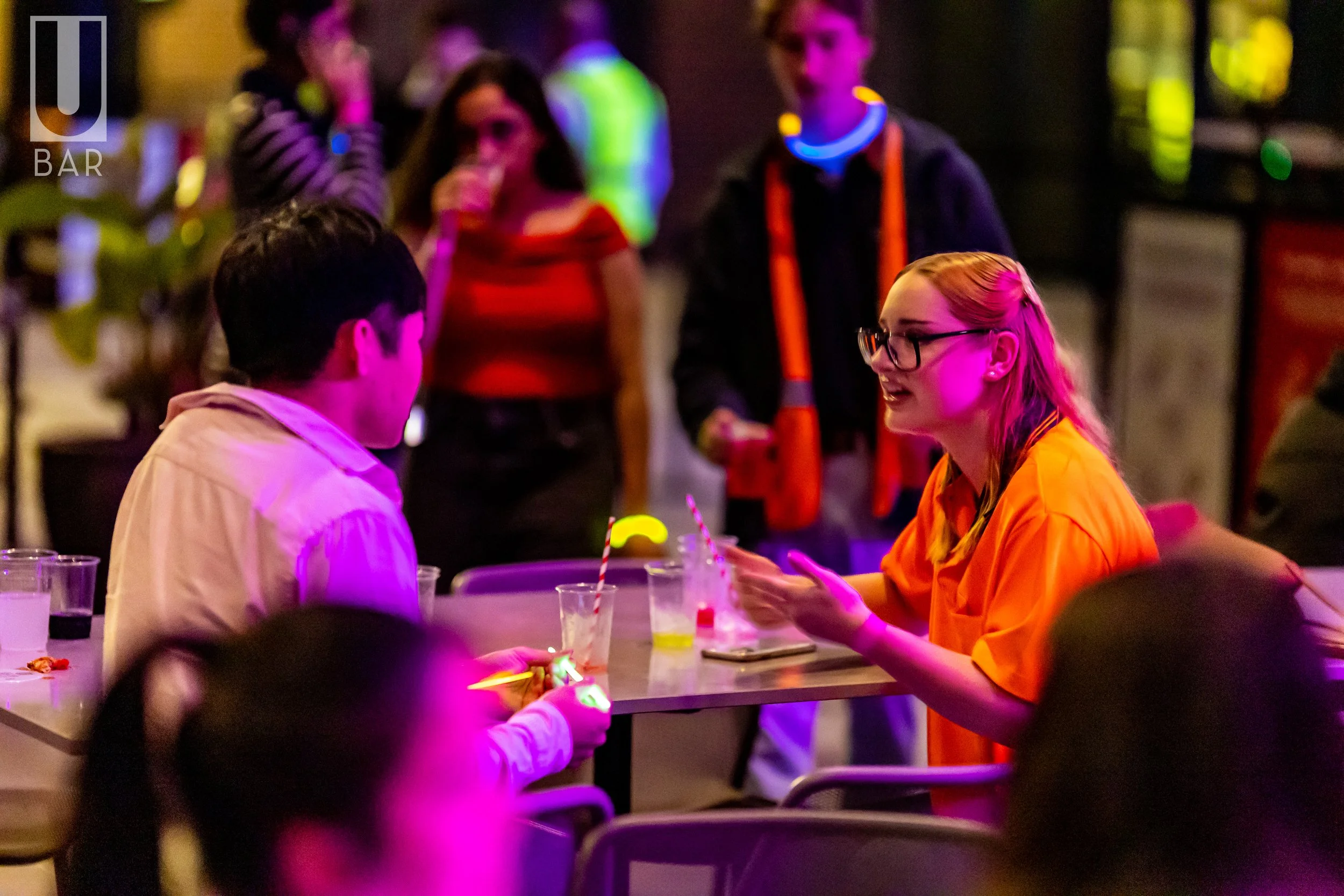 People socializing at a bar or nightclub with colorful lighting, some wearing glow necklaces, engaging in conversation, and holding drinks.