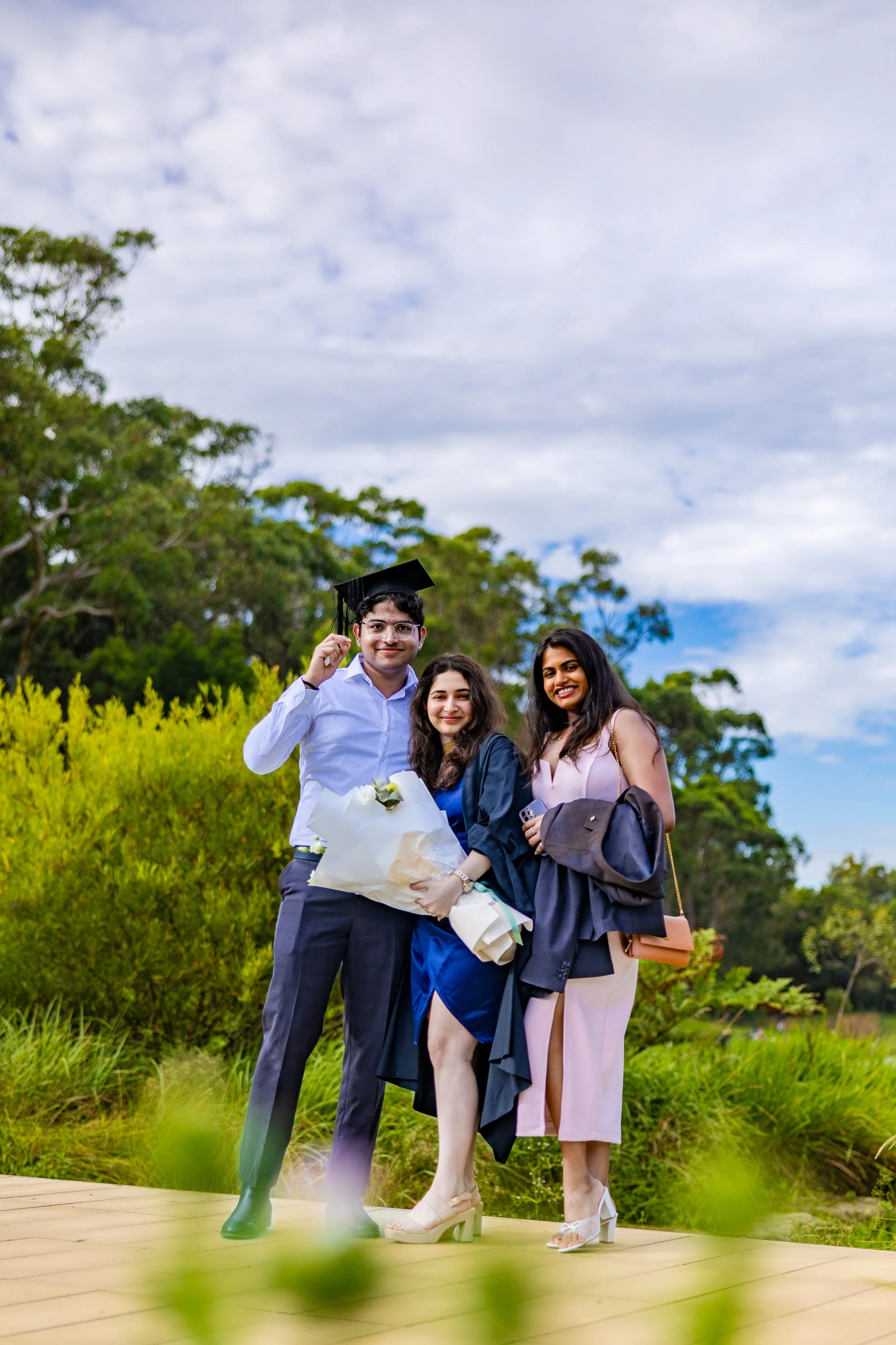 Group of three friends celebrating a graduation outdoors, with one person in a graduation cap and gown holding a bouquet of flowers, smiling, on a wooden platform with green foliage and trees in the background.
