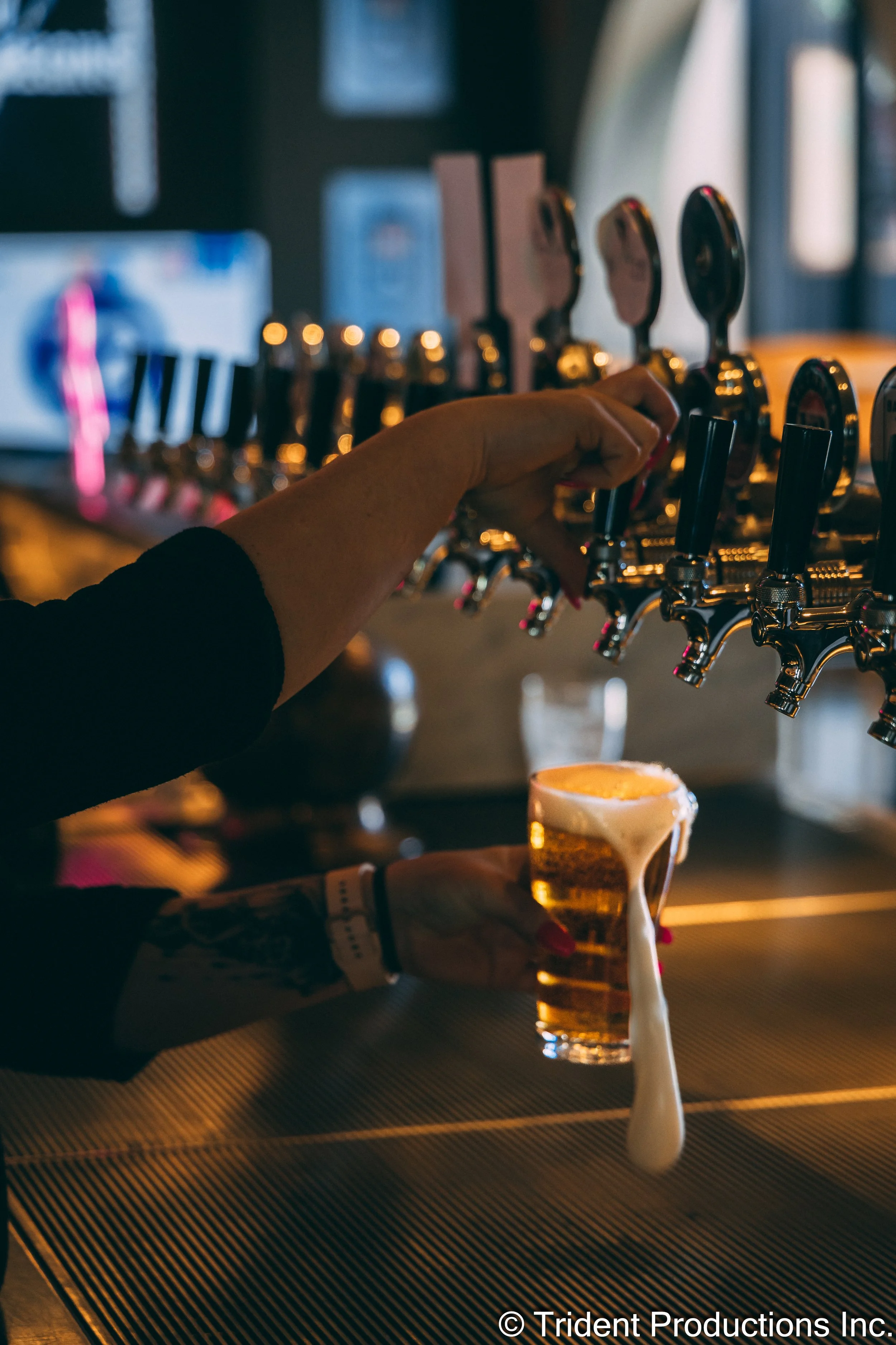 A person with tattoos on their arm is pouring beer from a tap into a glass, which overflows with foam, in a bar or pub setting.