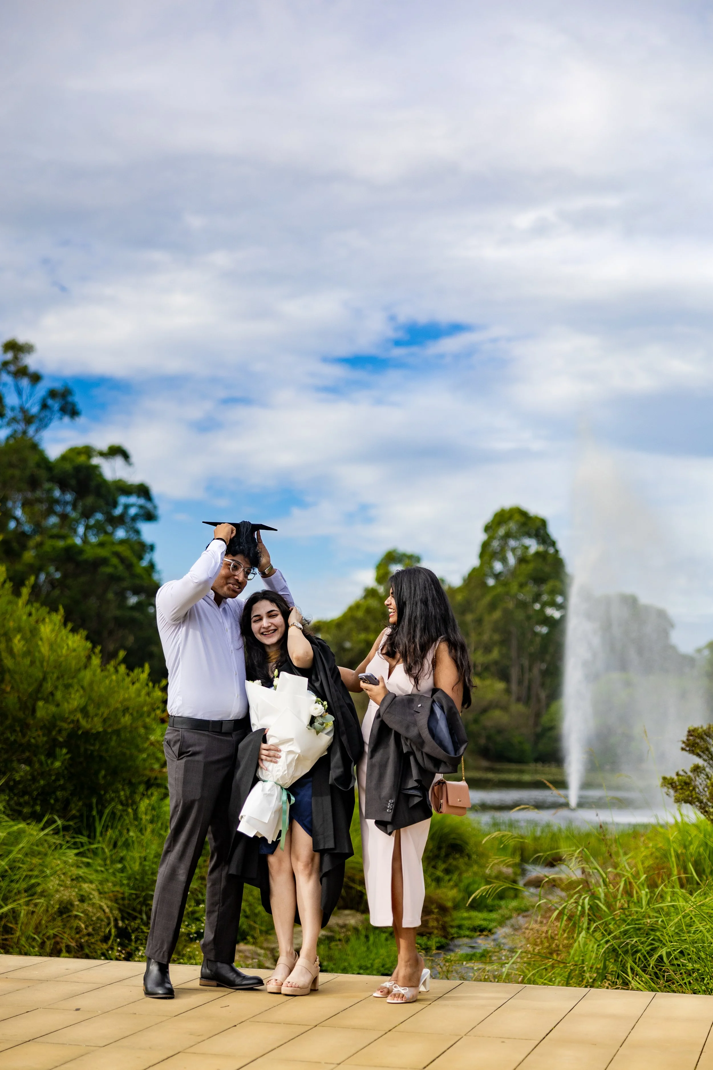 A graduate in cap and gown with a bouquet, celebrating with two friends outdoors near a fountain and greenery.
