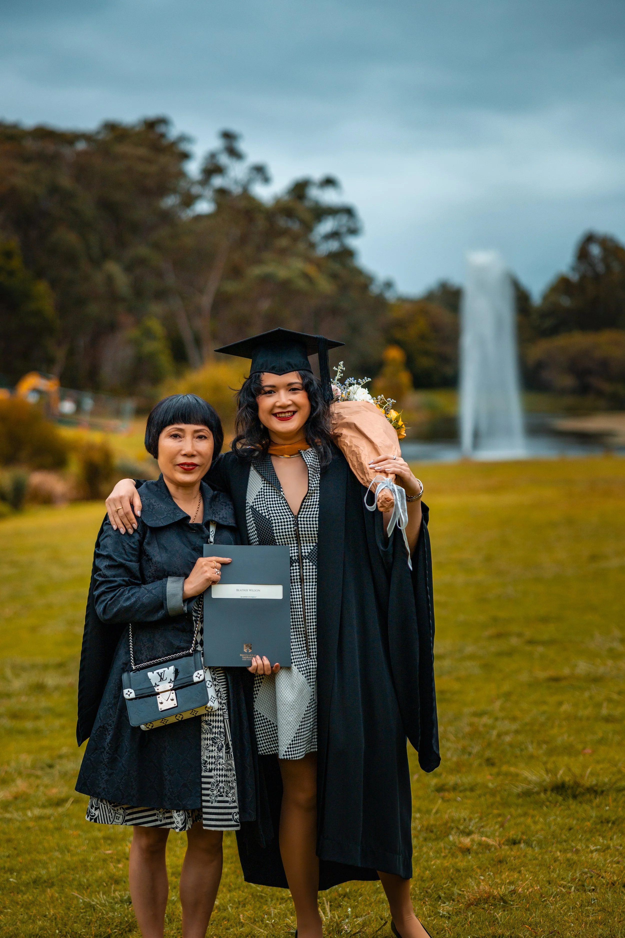 A woman in a graduation cap and gown holding a bouquet of flowers standing outdoors with another woman, both smiling, with a fountain and trees in the background.