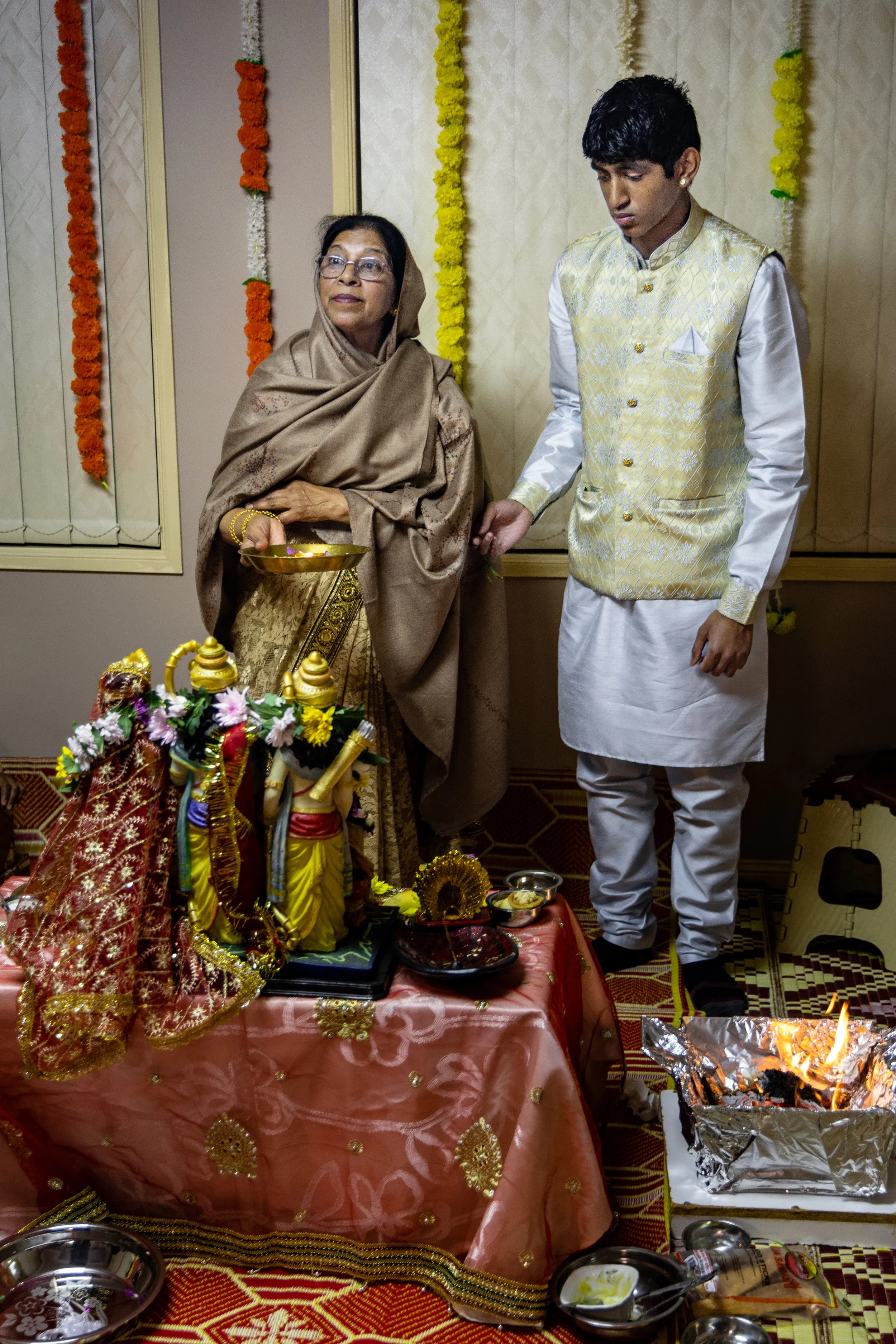 A woman and a young man participating in a traditional Hindu religious ceremony. The woman is holding a plate and wearing a brown shawl, while the young man is dressed in traditional Indian attire. They are standing in front of a decorated altar with