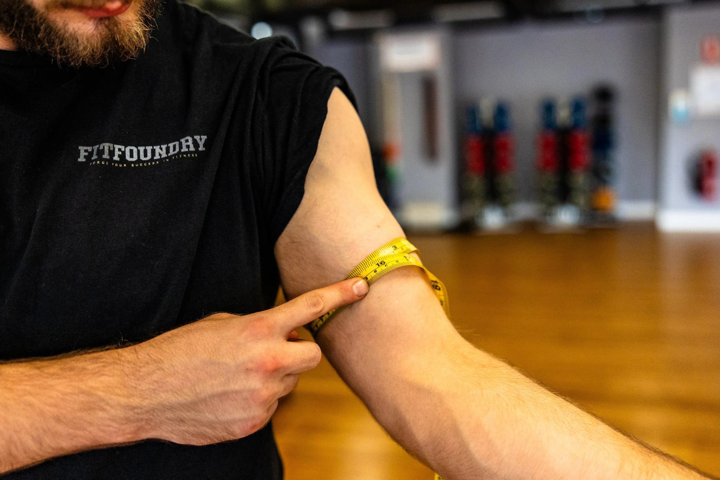 A man wearing a black shirt with the logo 'FitFoundry' measures his upper arm with a yellow measuring tape in a gym or fitness center.
