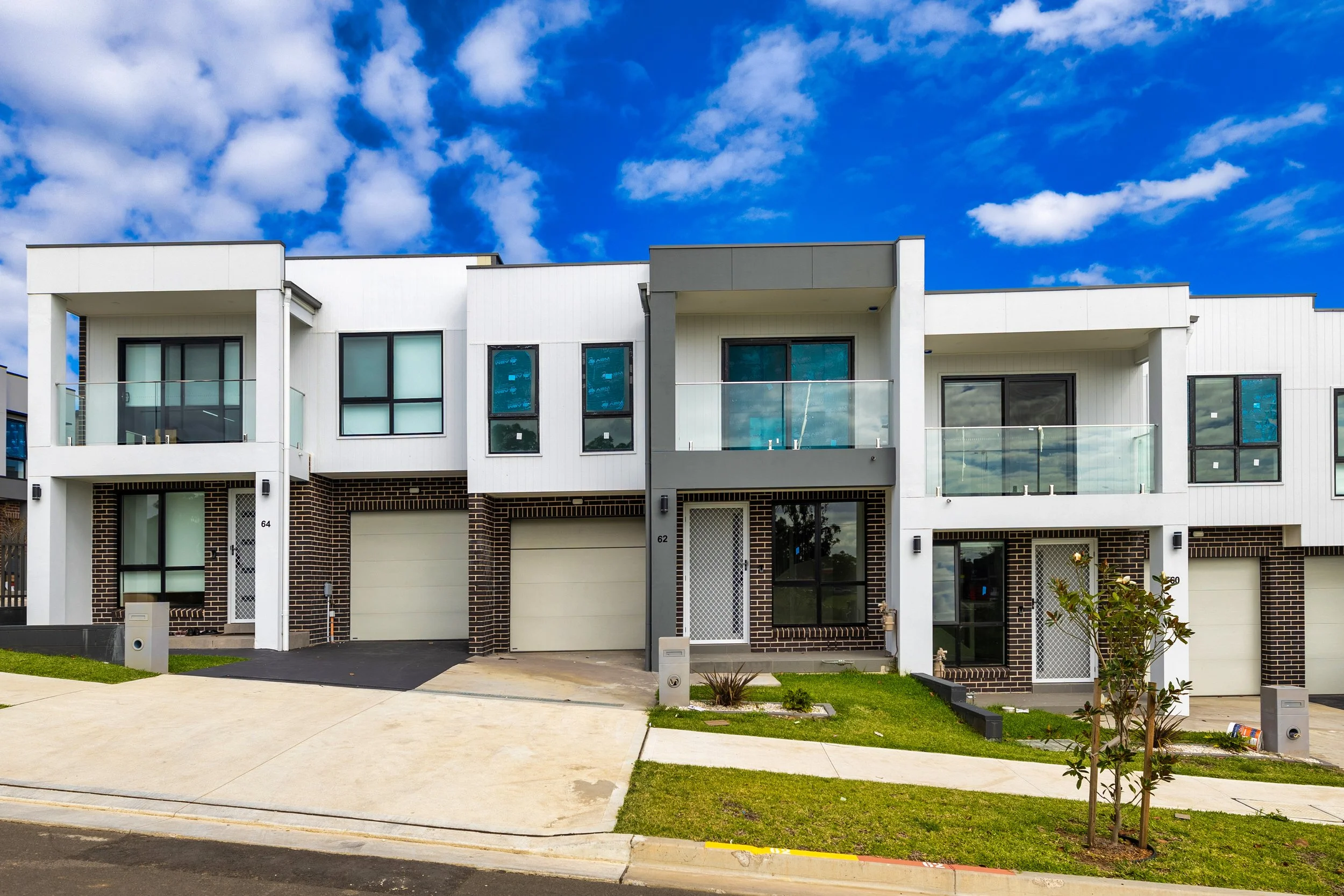 Modern townhouses with black, white, and brick exteriors, small lawns, and driveways in front, under a partly cloudy blue sky.