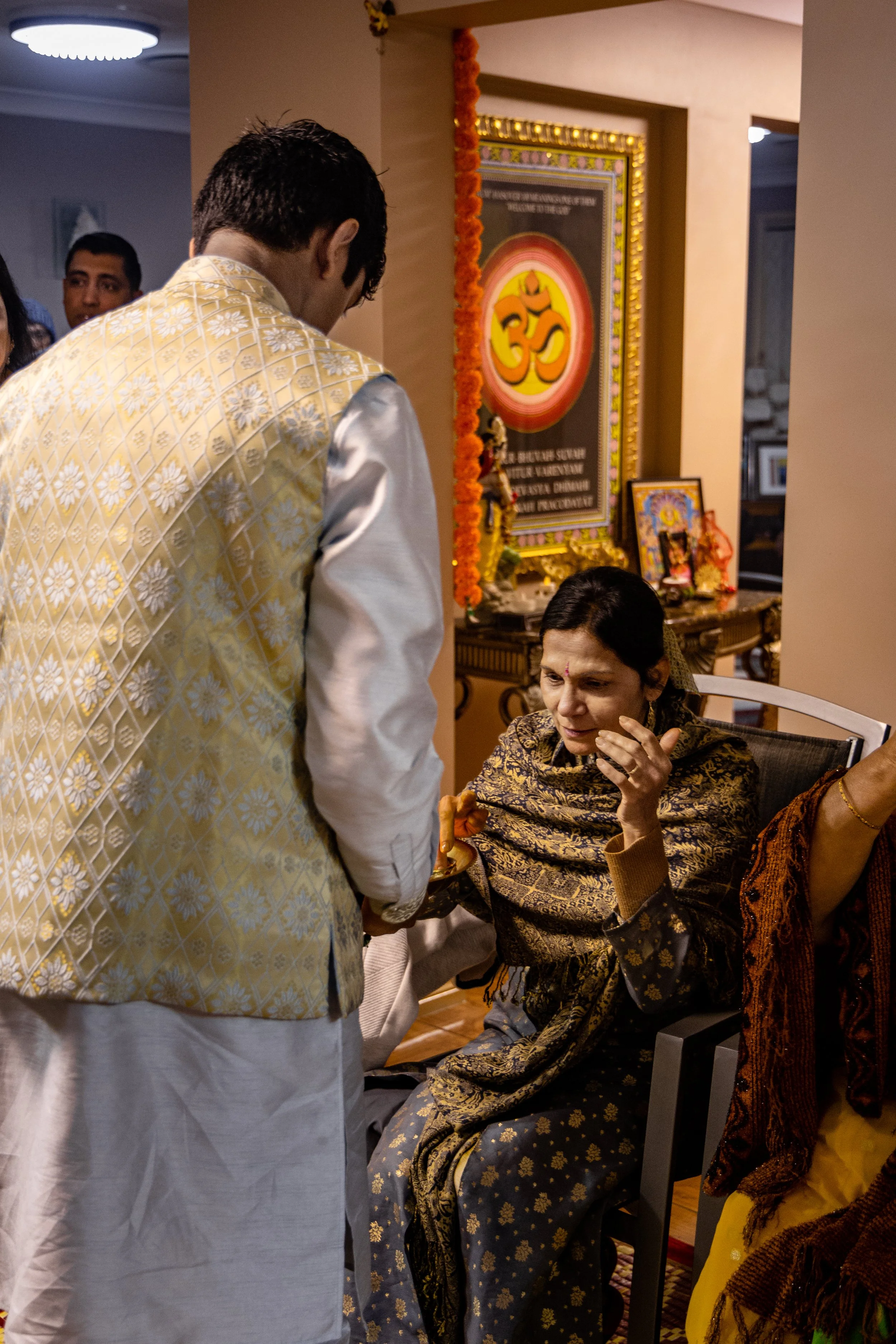 An elderly woman sitting on a chair, dressed in traditional Indian attire, receiving a plate of food from a man in a festive setting decorated for a religious celebration, with a framed religious symbol on the wall behind them.