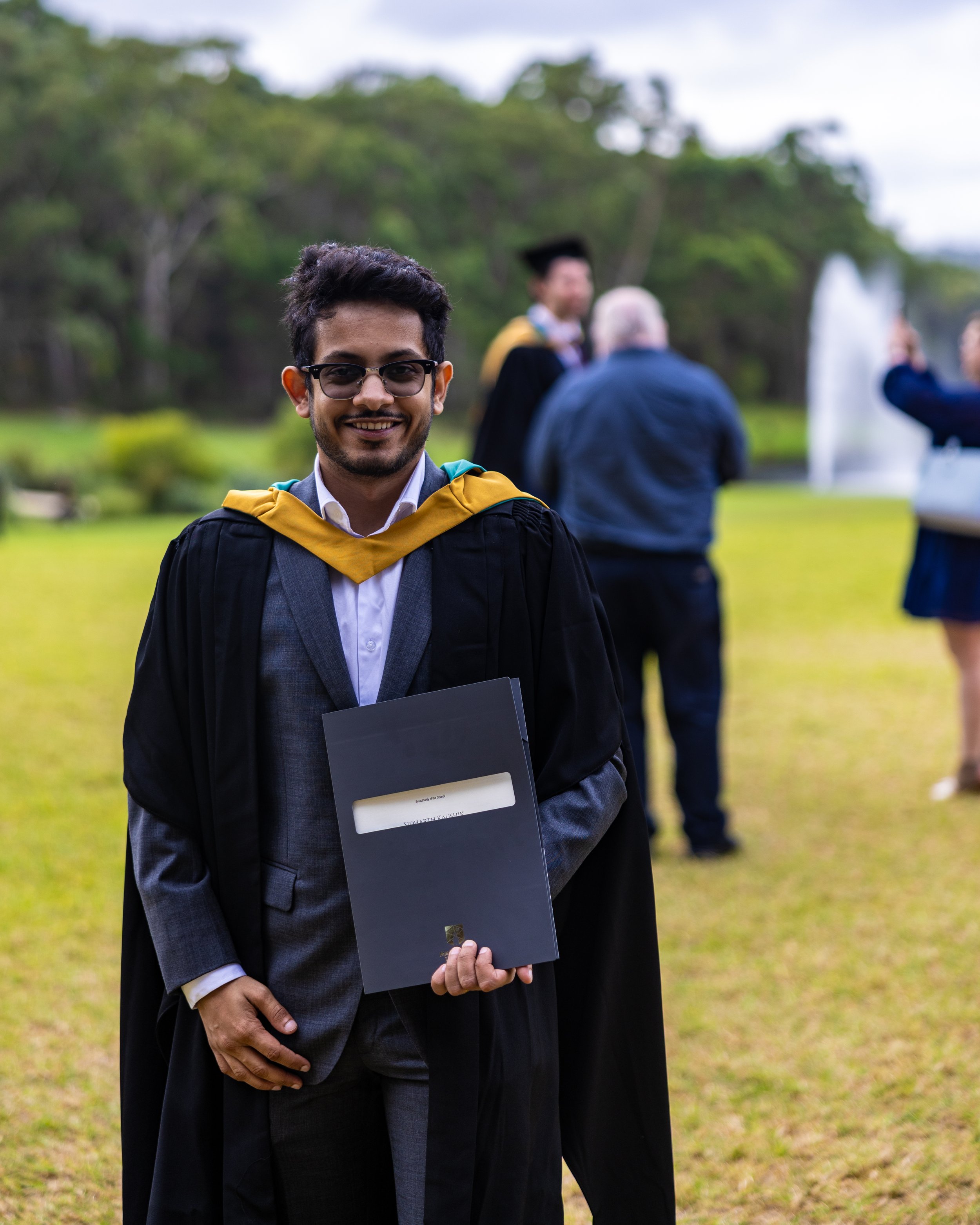 A young man in graduation attire, holding a diploma folder, smiling outdoors on graduation day with other graduates and attendees in the background.