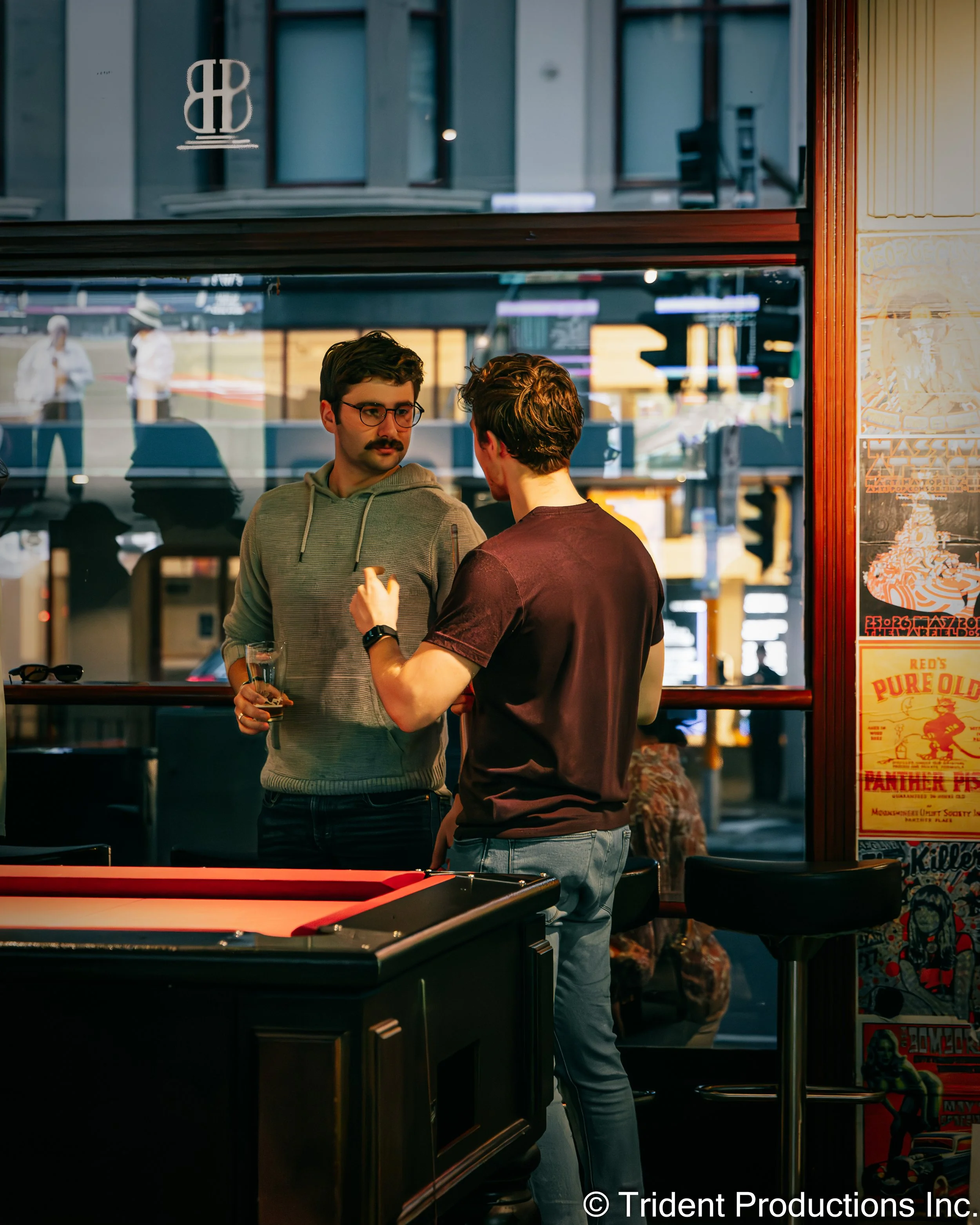 Two men having a conversation in a bar, one holding a glass of beer. They are standing near a pool table, with posters on the wall and a large window behind them reflecting outside city streets.