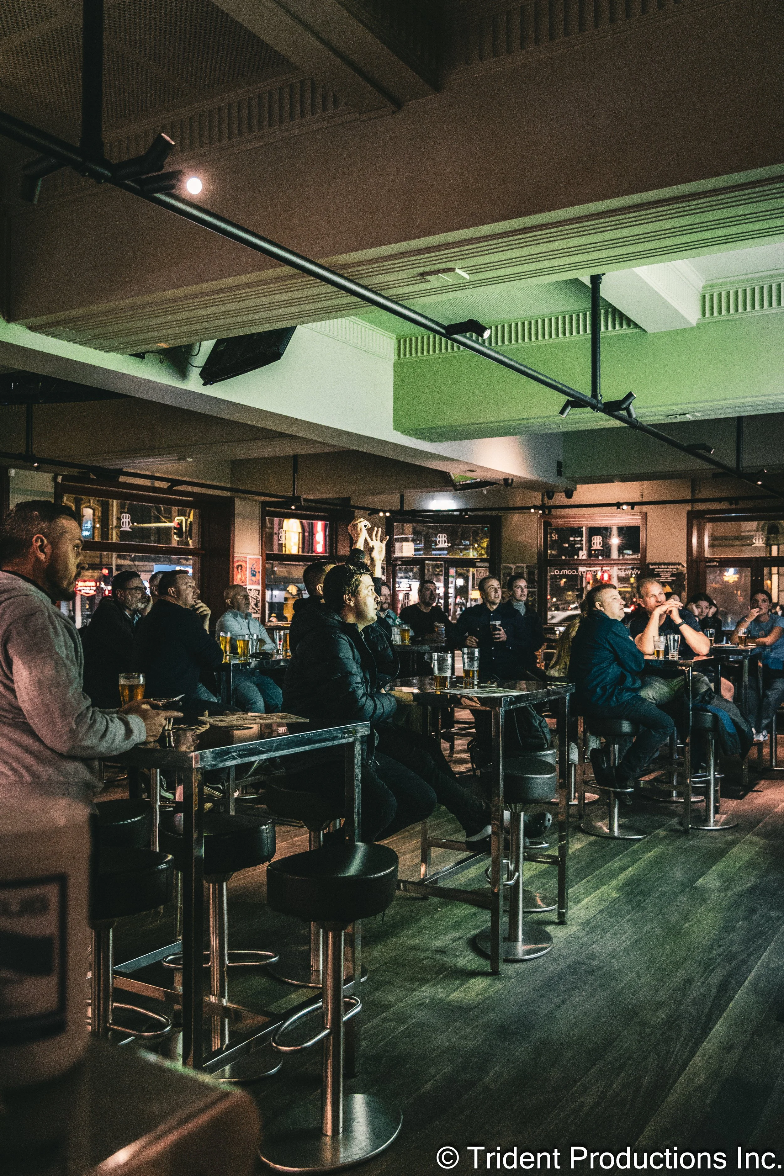 People watching a sports game on a big screen in a dimly lit bar or pub, seated at tables with drinks.
