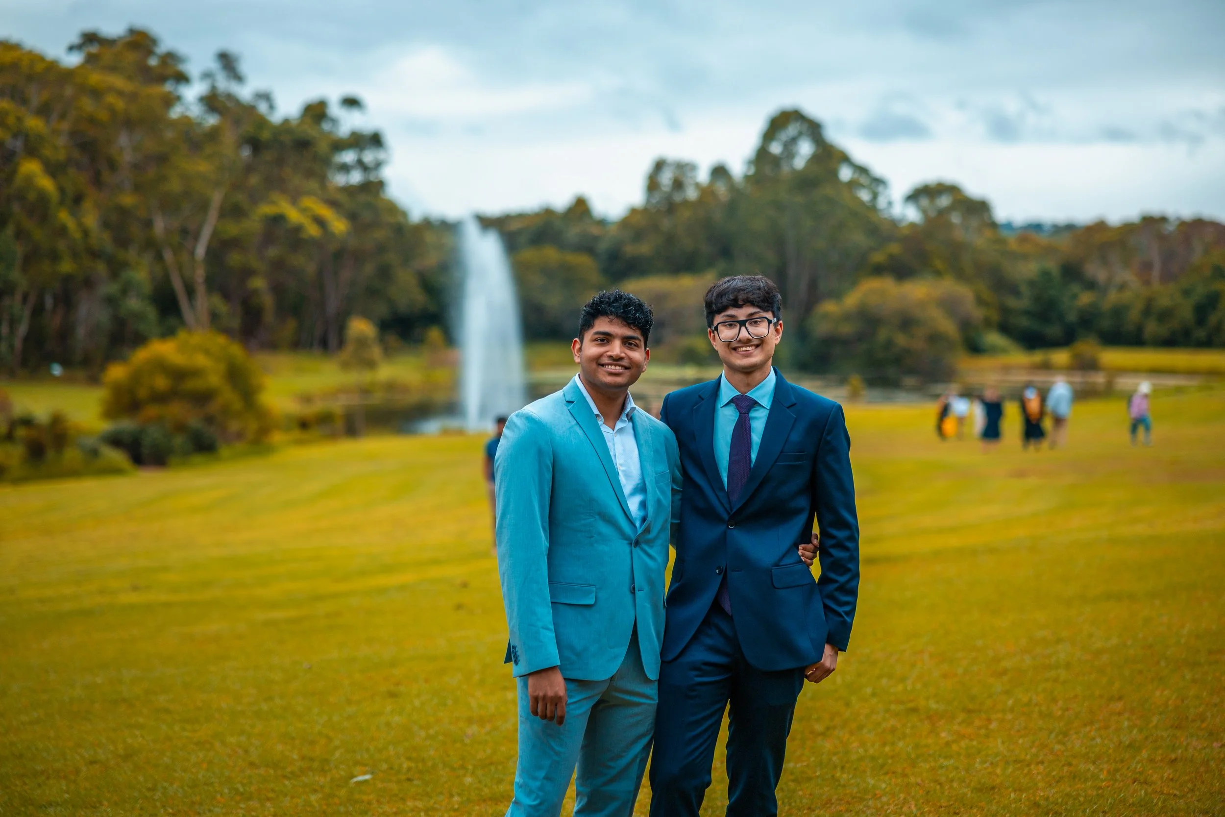 Two young men in suits standing outdoors on a grassy field, smiling at the camera with a fountain and trees in the background.