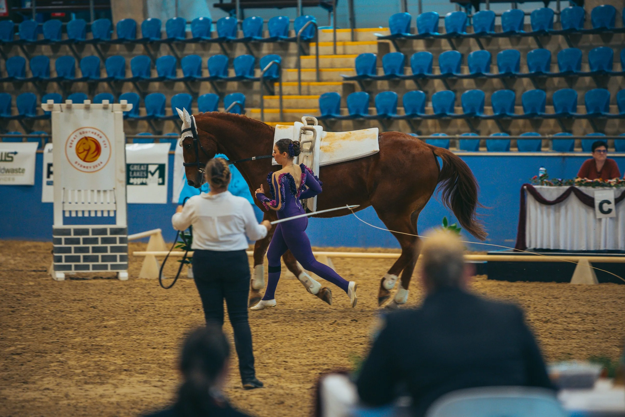 A horse with a saddle and bridle is being handled by a woman in a white jacket, while a female figure skater in a purple costume is walking beside it, in an indoor arena with blue seats and judges' tables.
