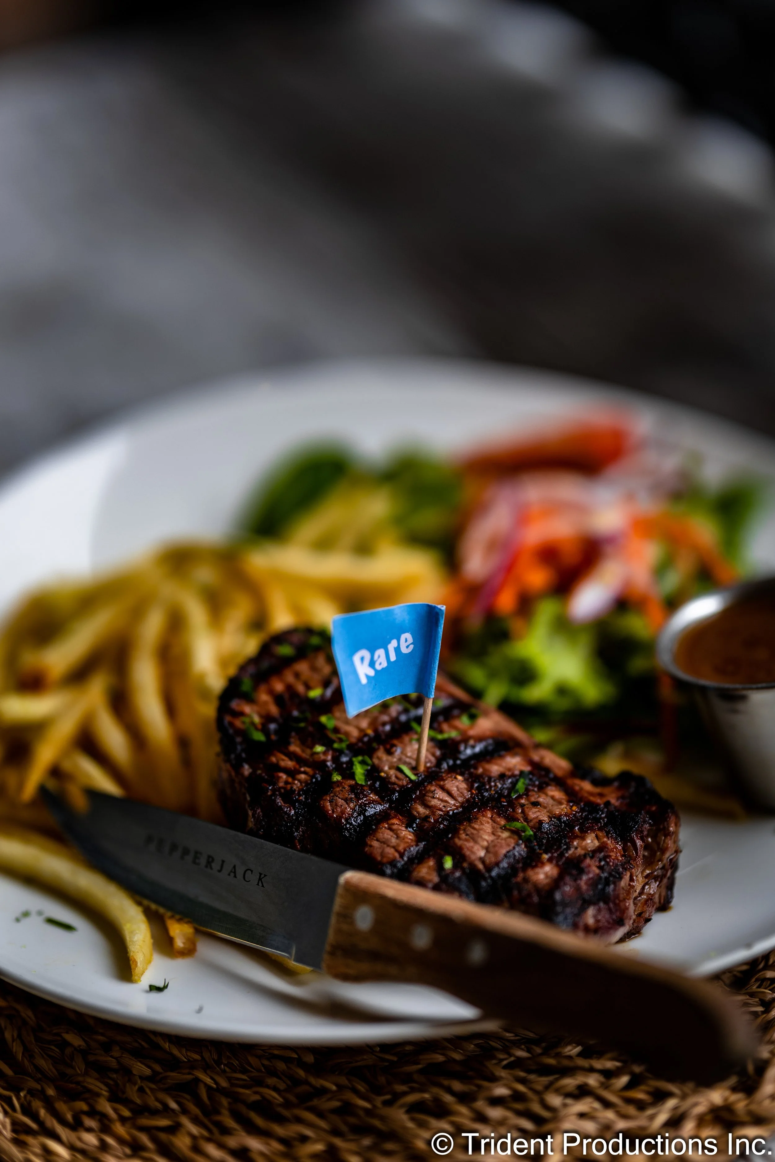 A grilled steak topped with chopped herbs on a white plate, accompanied by French fries, salad, and a small container of sauce, with a small blue flag labeled 'Rare' in the steak.