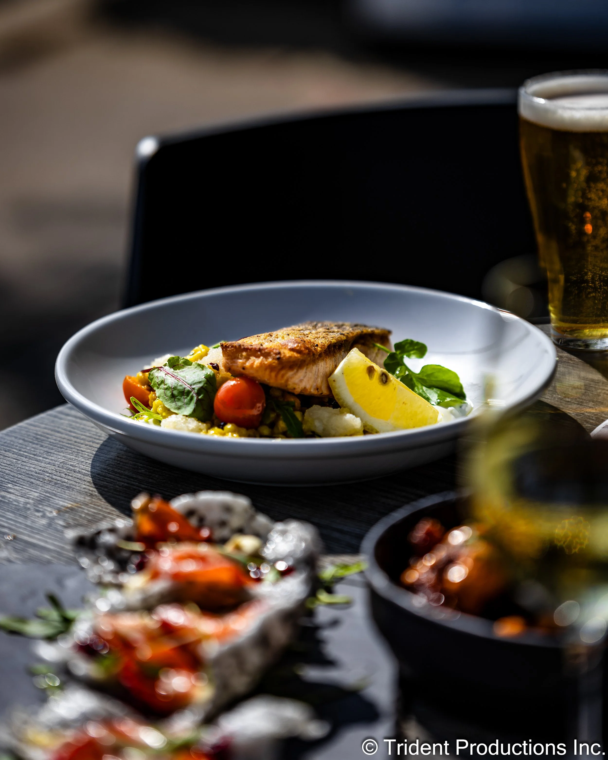 A plated dish of grilled fish with vegetables and lemon wedge, accompanied by a glass of beer on a dark wooden table.