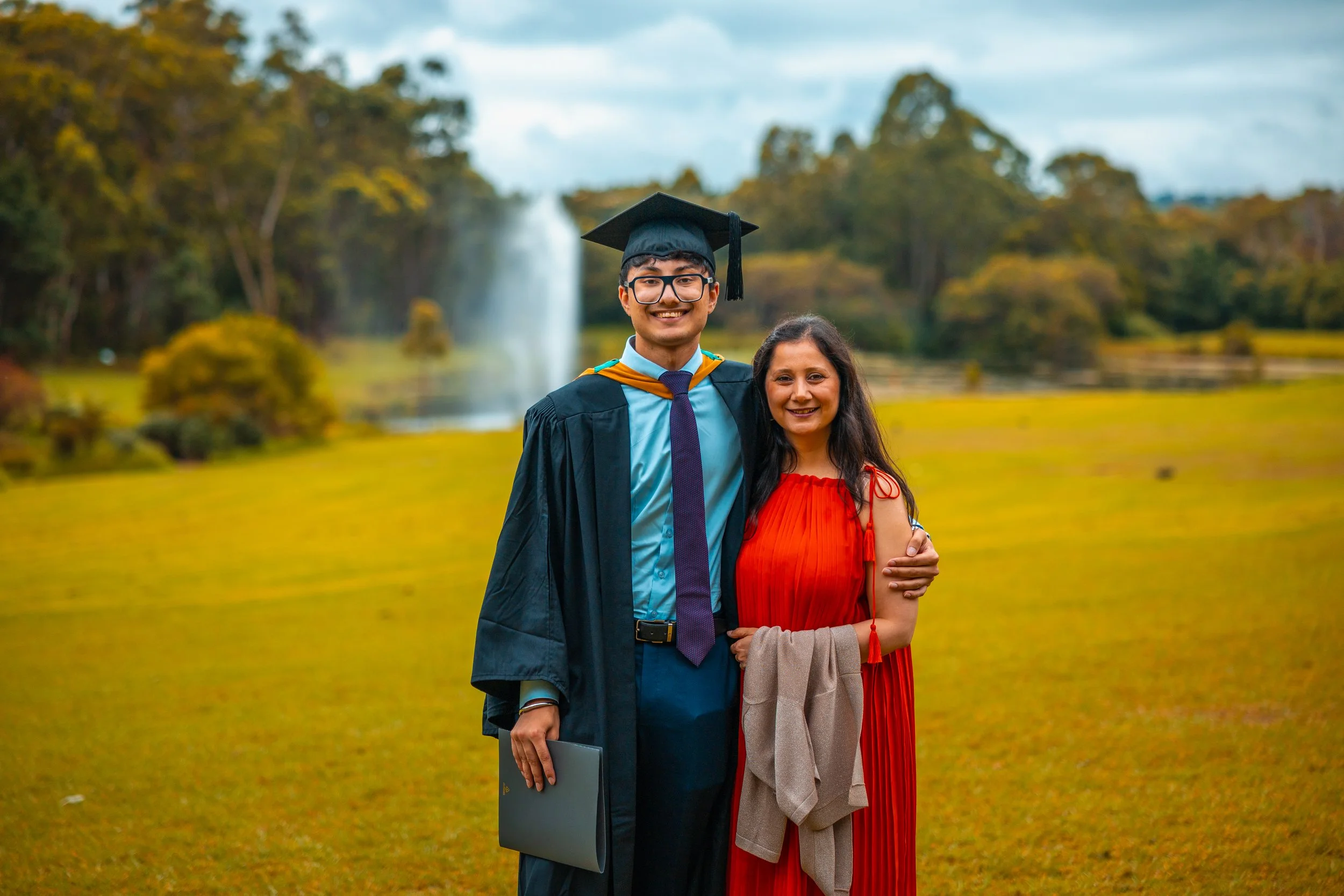 A young man in a graduation cap and gown standing next to a woman in a red dress, both smiling, outdoors near a pond with a fountain in the background.