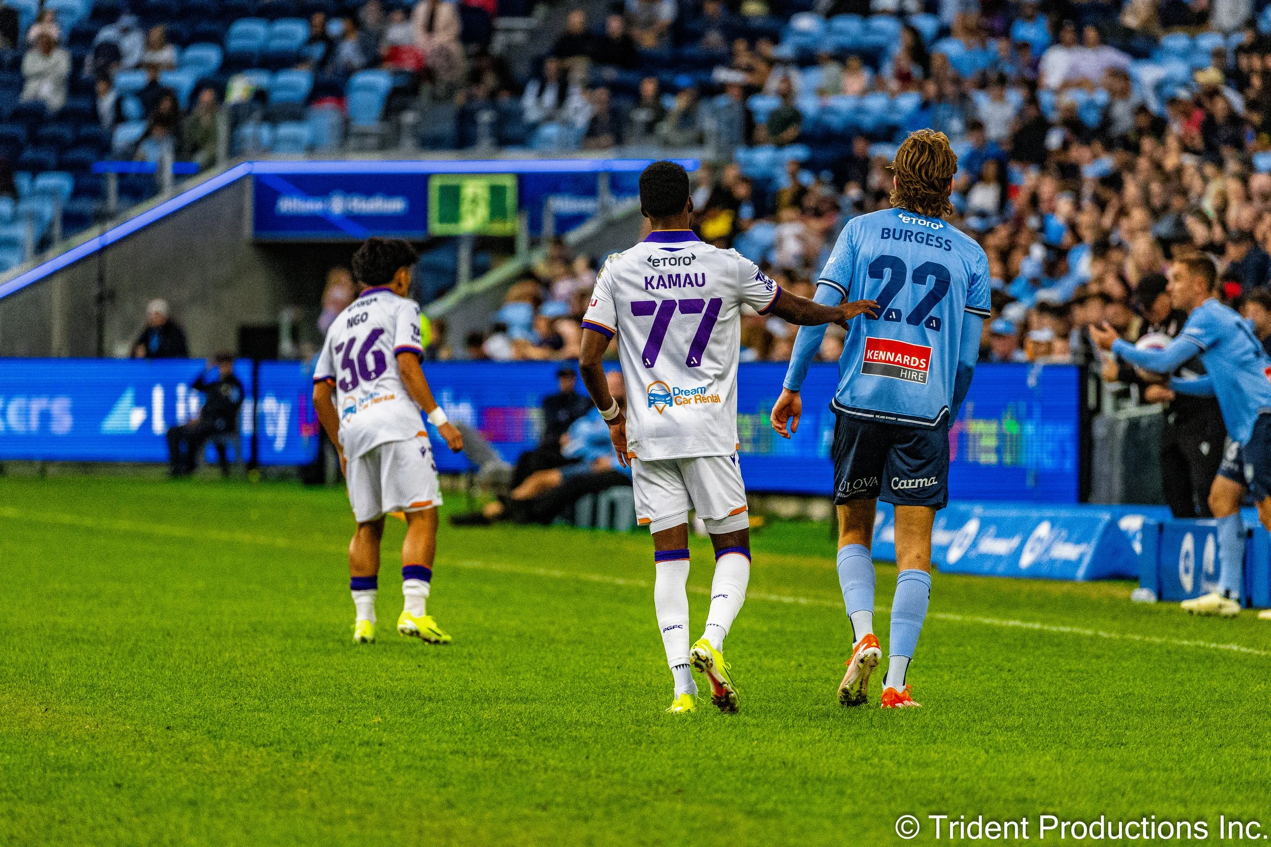 Two soccer players walk on the field, with one player placing his hand on the back of the other. The players are wearing blue and white uniforms with various sponsor logos, and there is a crowd of spectators in the stands.