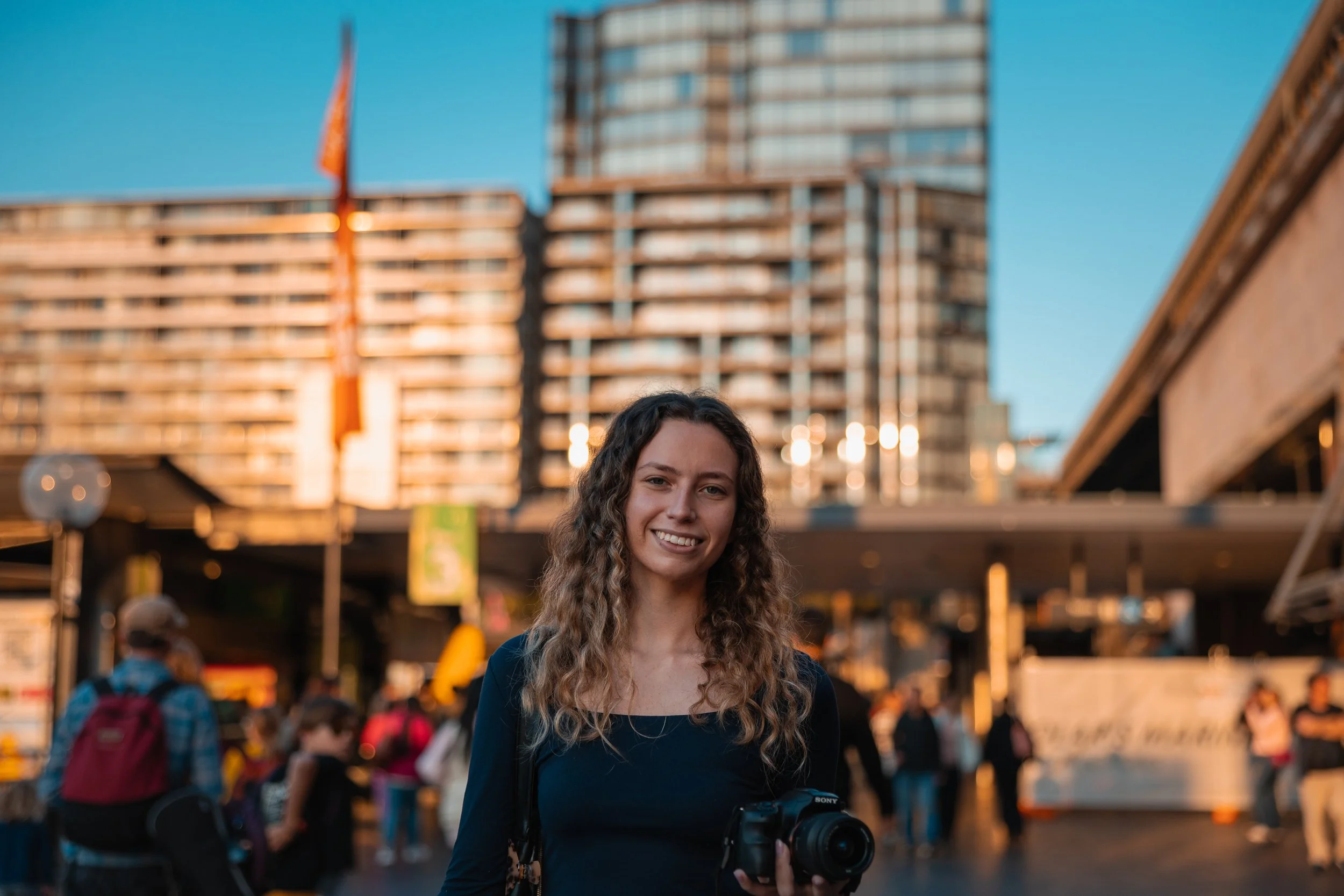 Young woman with curly hair smiling at camera, holding a camera, with urban cityscape and buildings in the background during sunset.