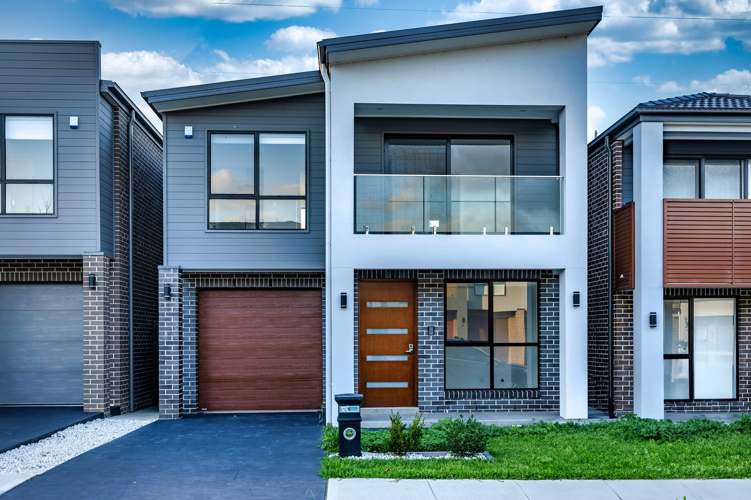 Front view of a modern two-story house with a brick and white exterior, large windows, and a wooden front door, situated between two similar houses with garages and well-maintained lawns.