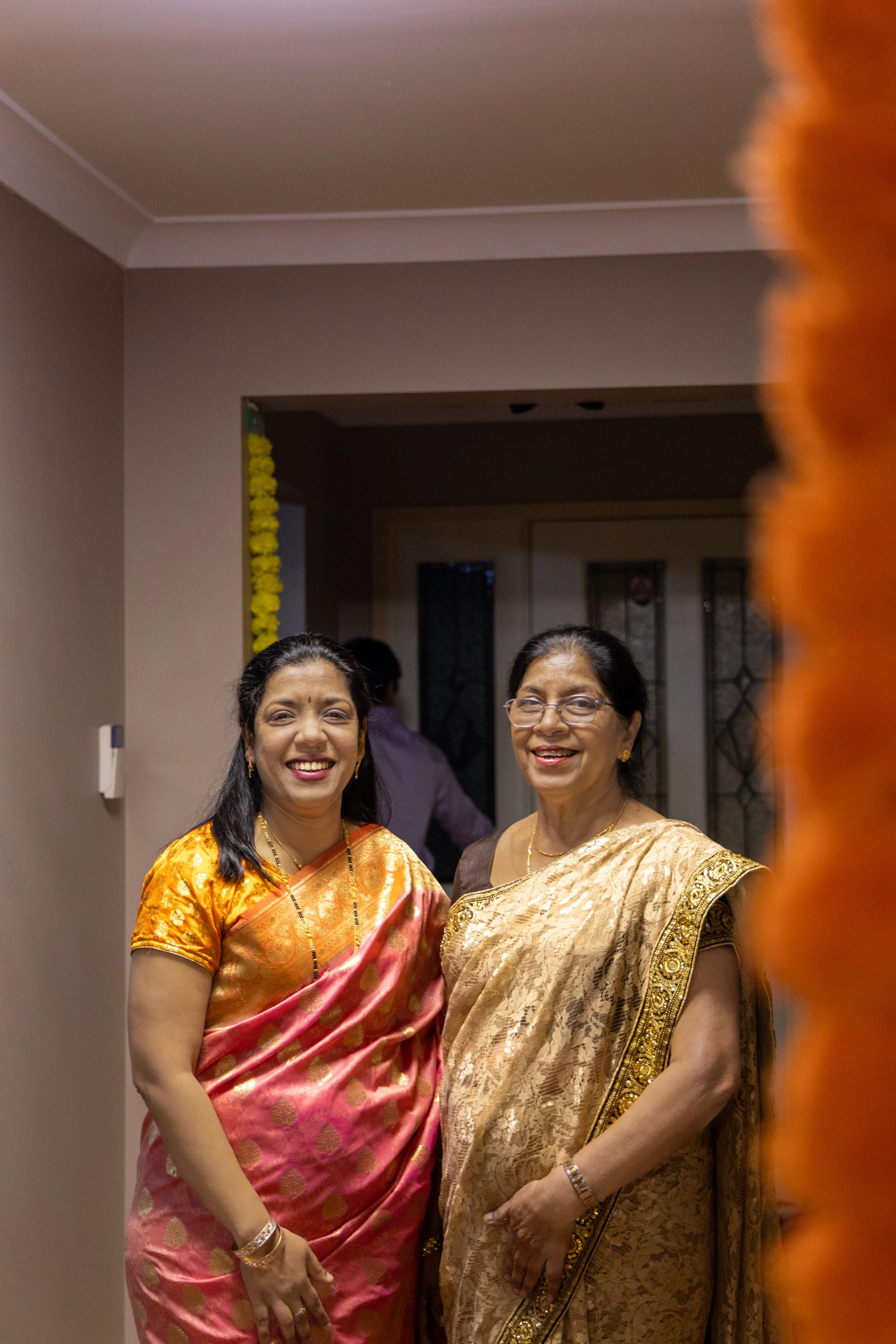 Two women wearing traditional Indian sarees smiling at a celebration or gathering indoors.