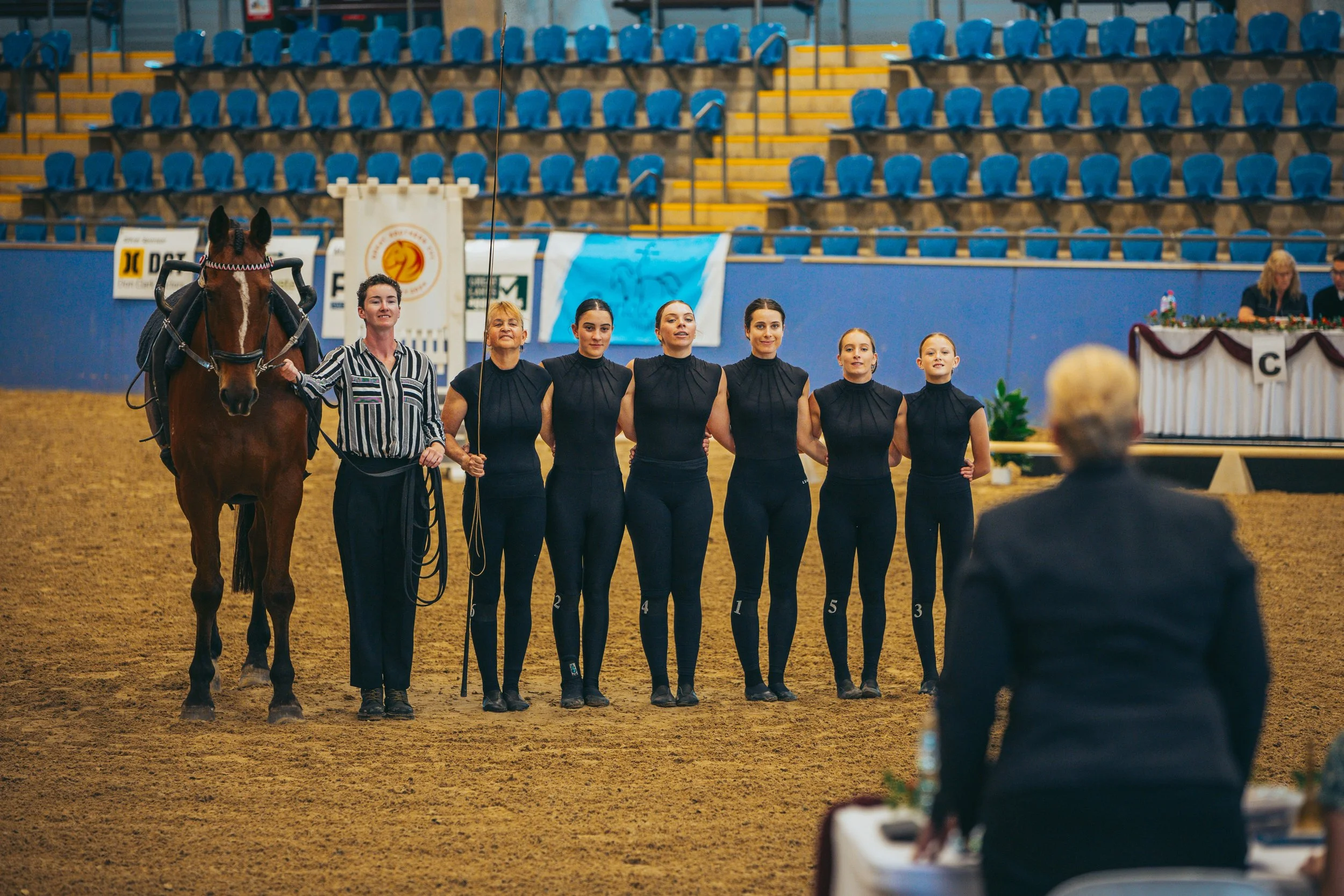 A group of six women in black outfits standing in a line with a woman holding a horse and a judge observing in the foreground at an indoor equestrian event.