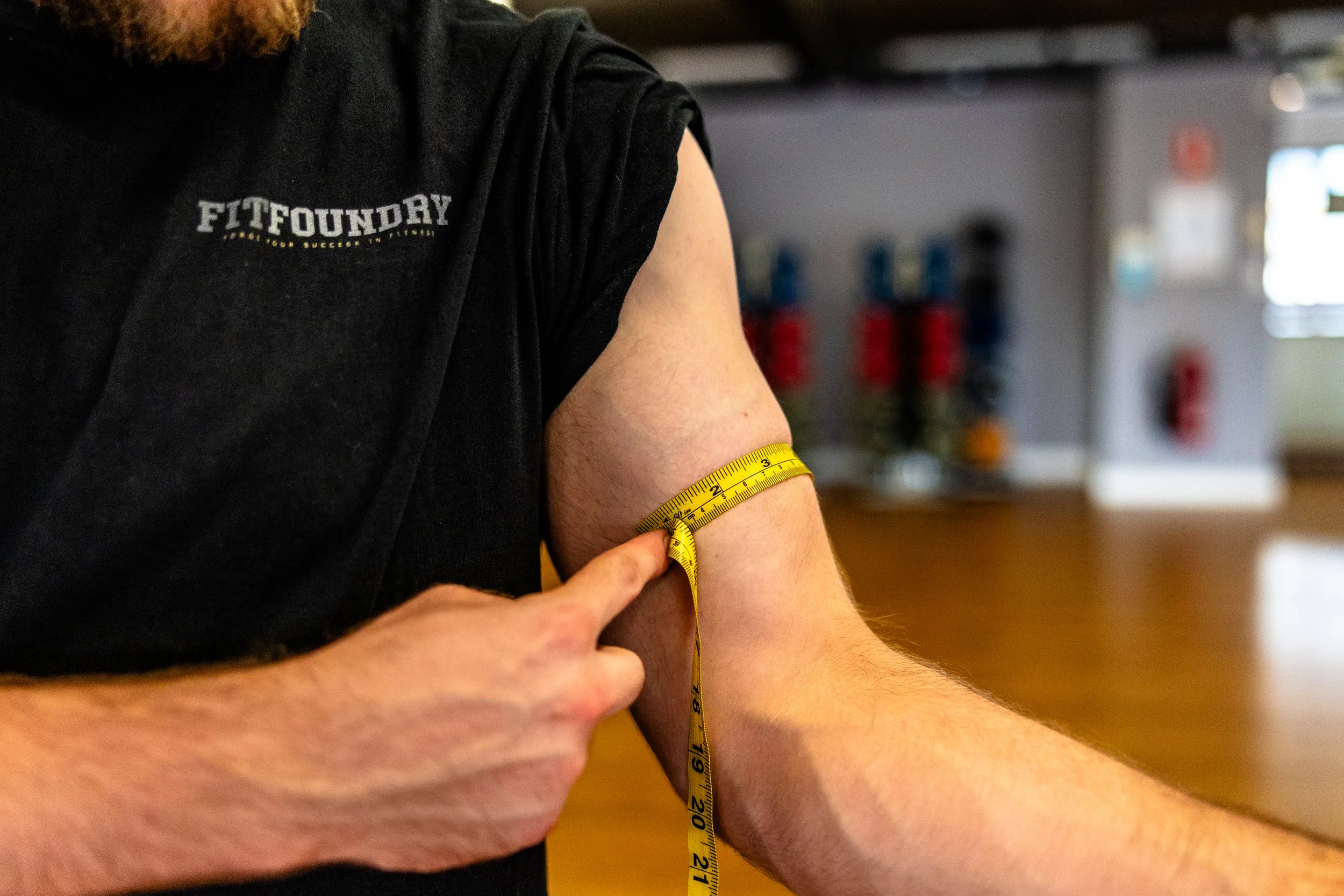 A person measuring their upper arm with a yellow measuring tape at a gym or fitness facility.