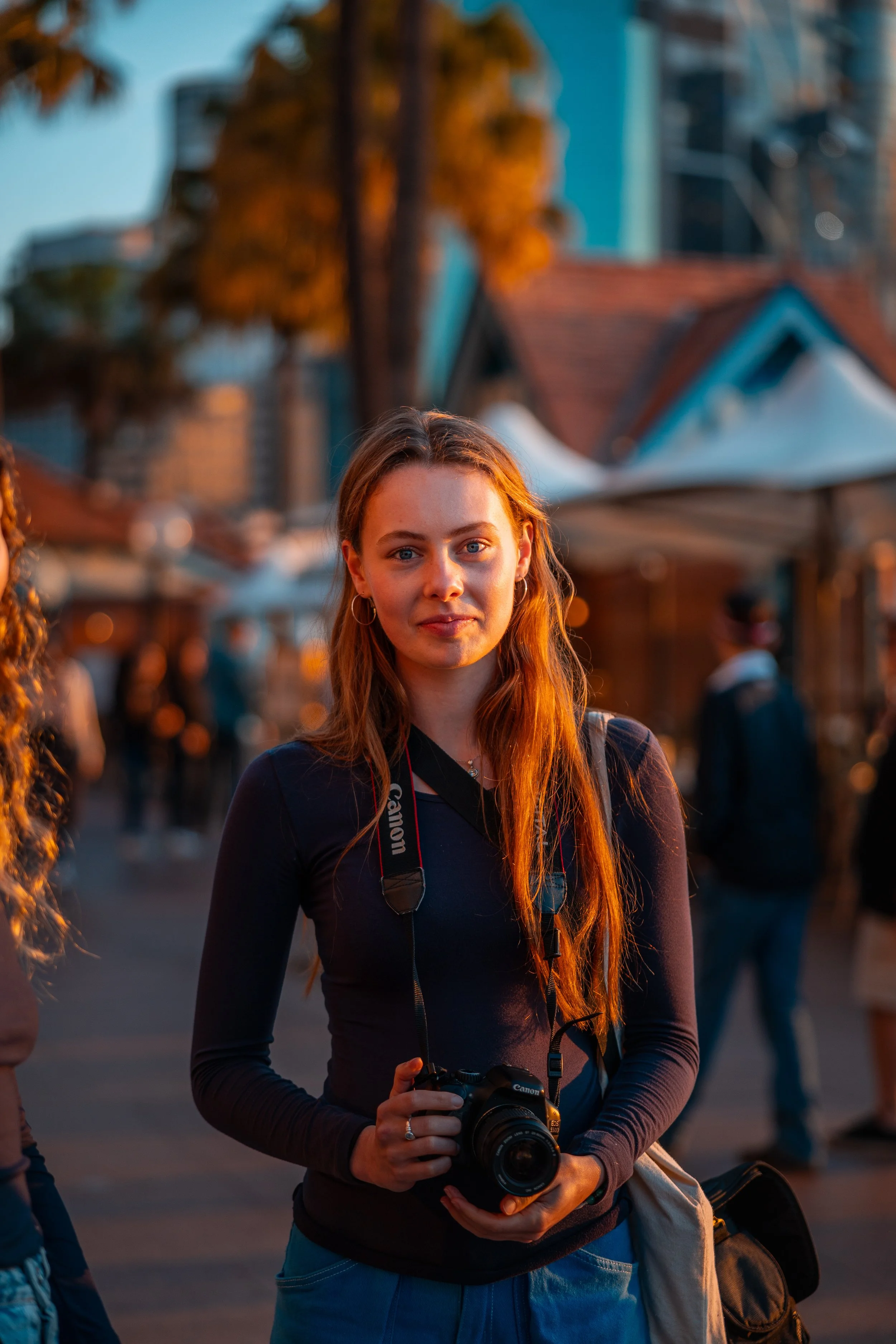 A young woman with long red hair holding a Canon camera, standing outdoors during sunset with a crowd and tents in the background.