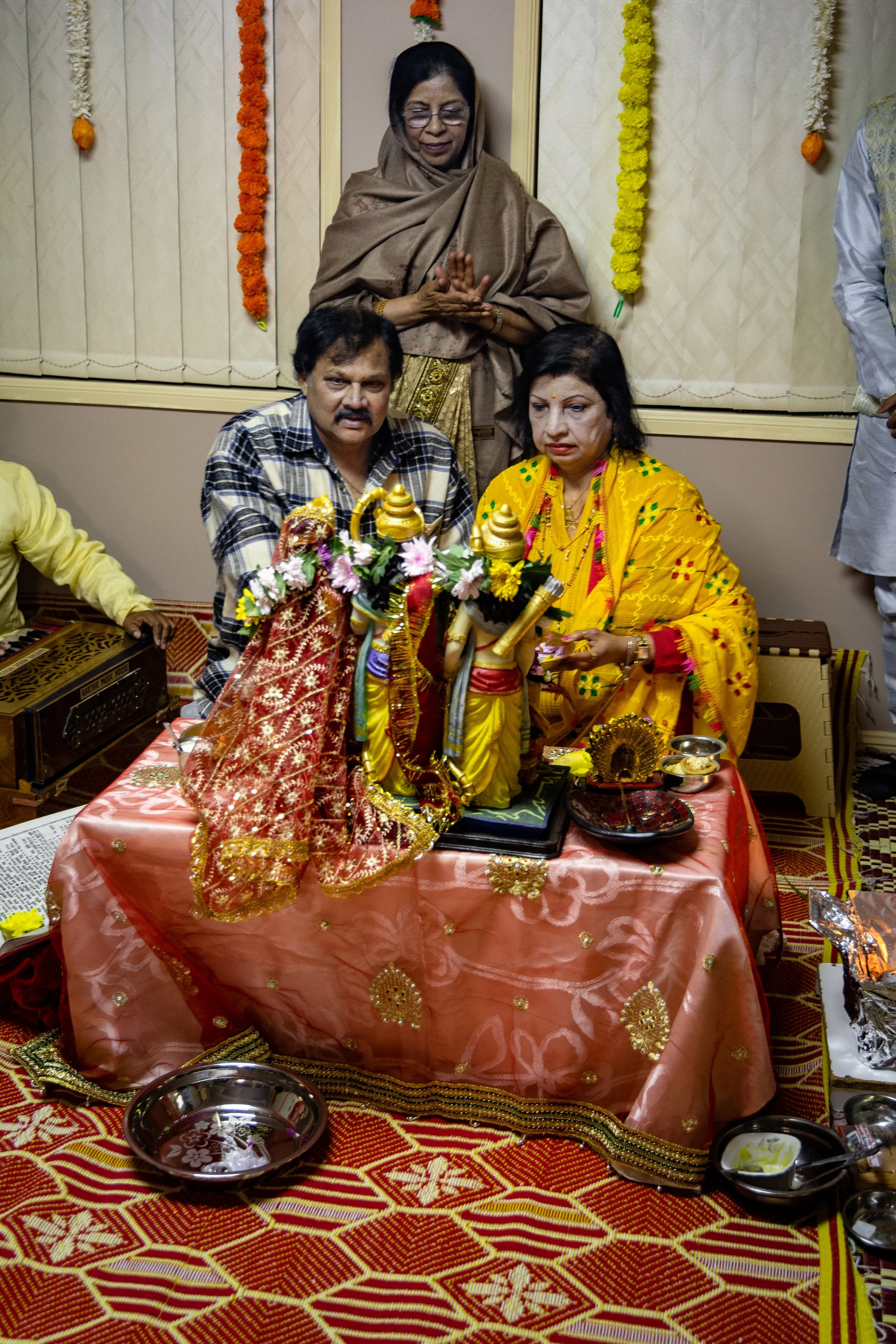 A Indian ceremony with a decorated table featuring Hindu idols, flowers, and offerings, with a woman in a yellow saree and a man in a checked shirt sitting at the table, and a woman in a brown shawl standing behind them.