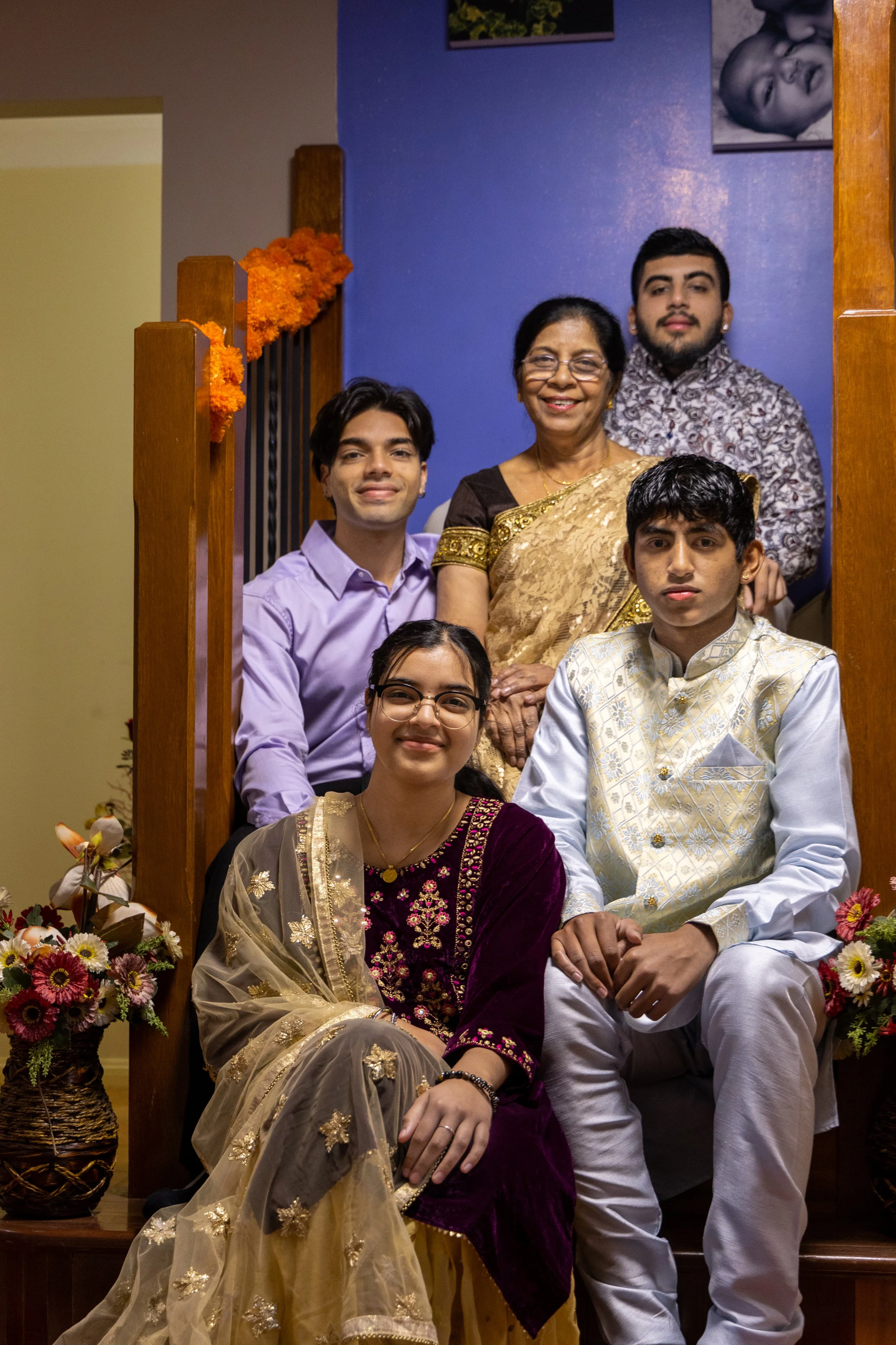 A multigenerational family of six poses on a staircase decorated with orange marigold garlands, wearing traditional Indian attire, in a home setting with framed artwork on the wall.