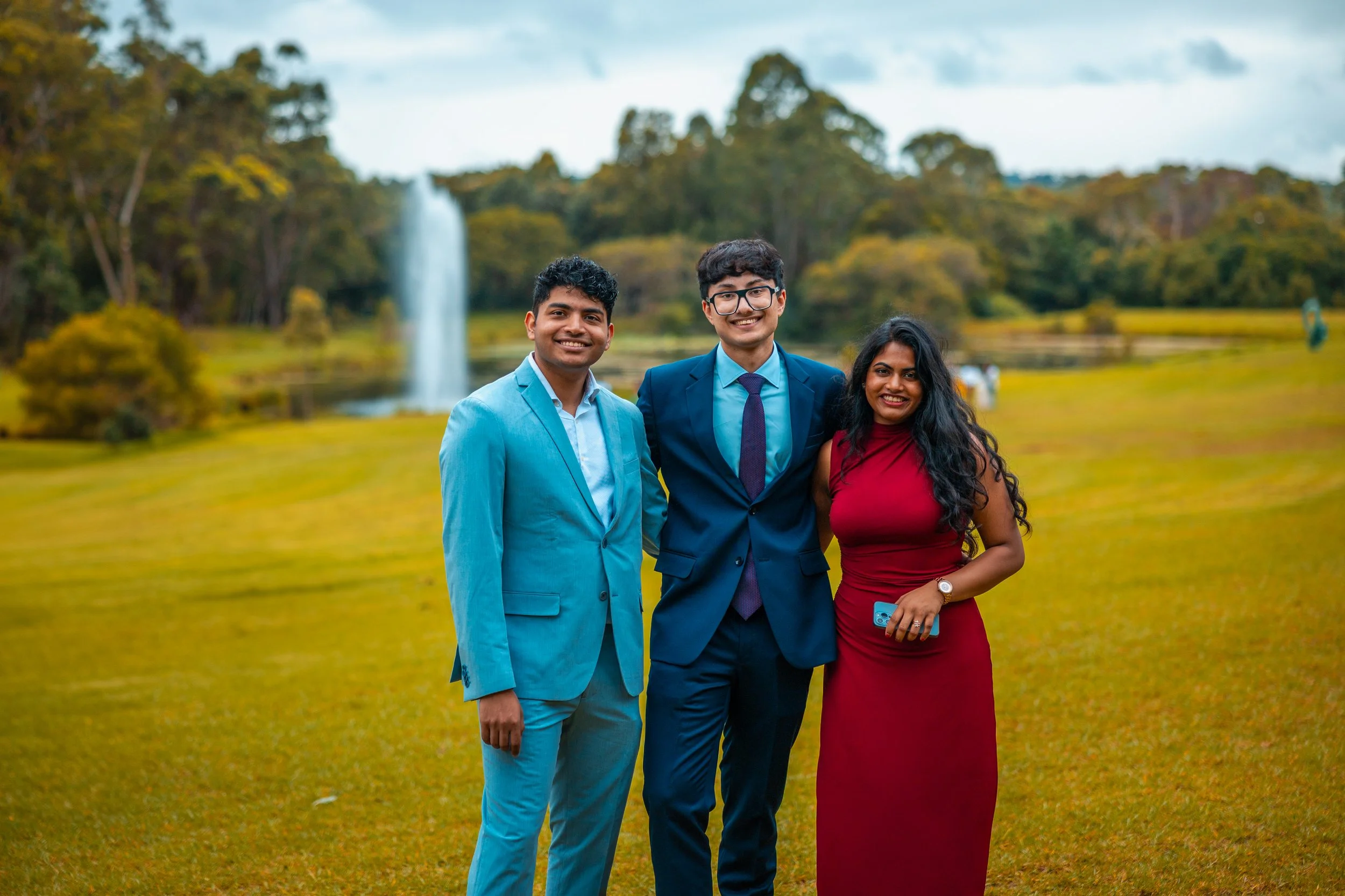 Three young adults standing outdoors on a grassy area with trees and a waterfall in the background. The group includes two men in suits and a woman in a red dress, all smiling.