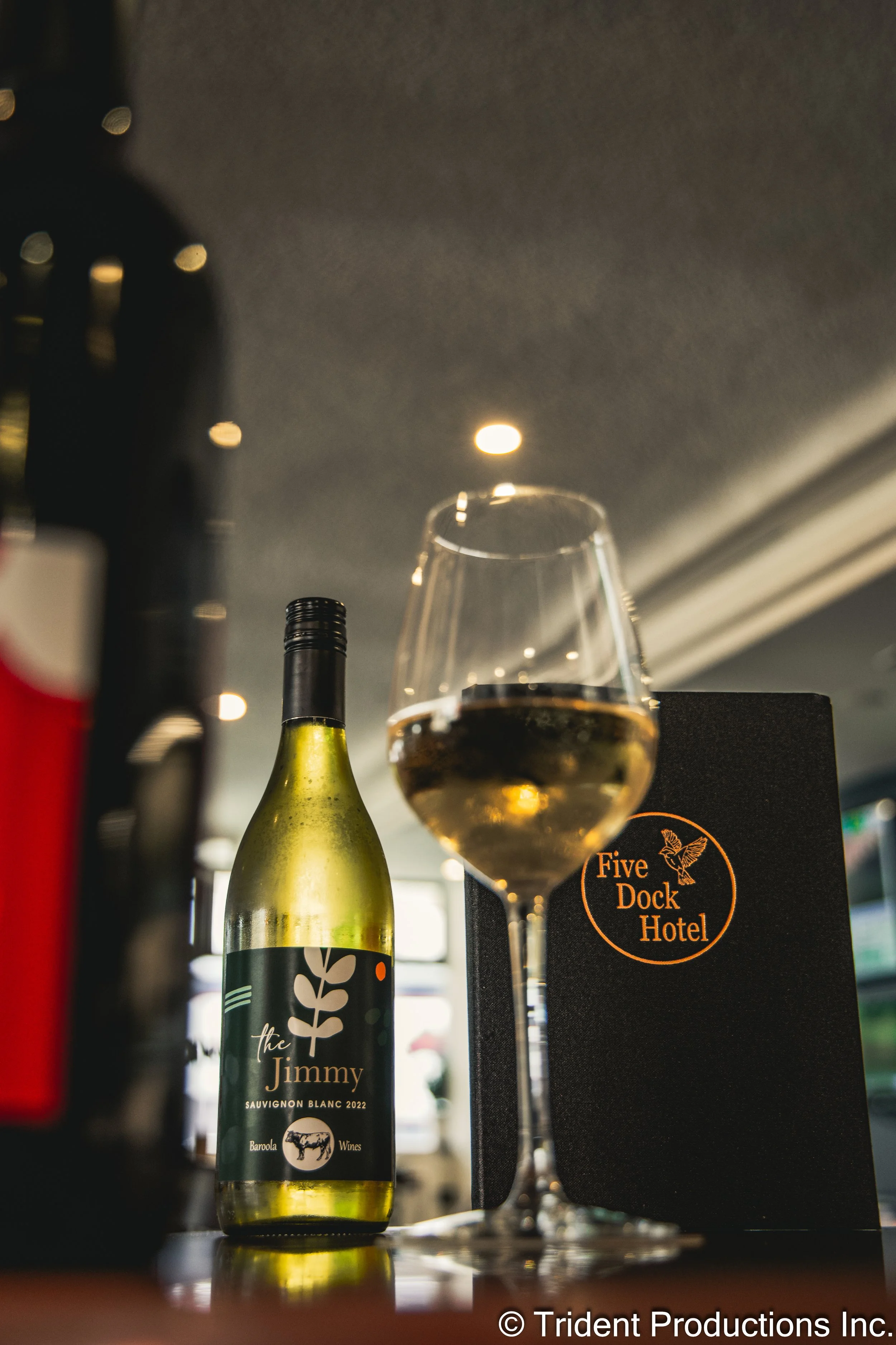 A bottle of white wine labeled 'the Jimmy Sauvignon Blanc 2022' stands next to a glass of white wine, on a bar counter in front of a black menu from the Five Dock Hotel.