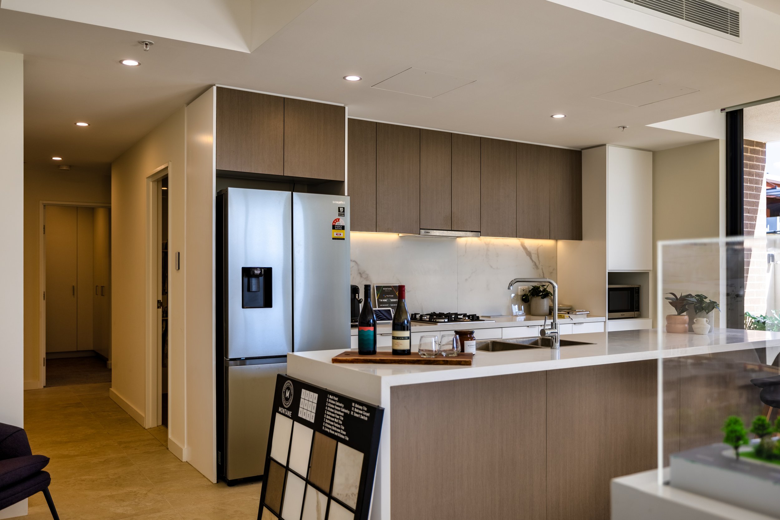 Modern kitchen with stainless steel refrigerator, wooden cabinets, marble backsplash, white countertop, and small potted plant.