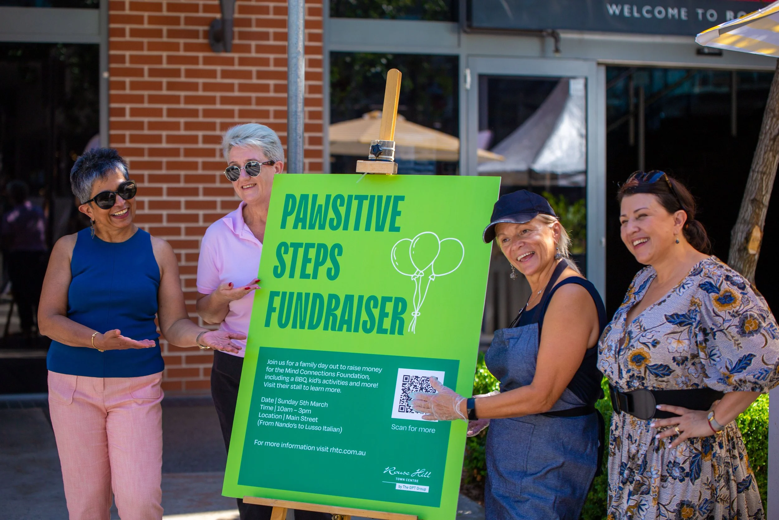 Four women standing outdoors at a Pawsitive Steps Fundraiser event, smiling and talking. One woman is holding a green sign with event details, including a QR code. The background features a brick building and trees.