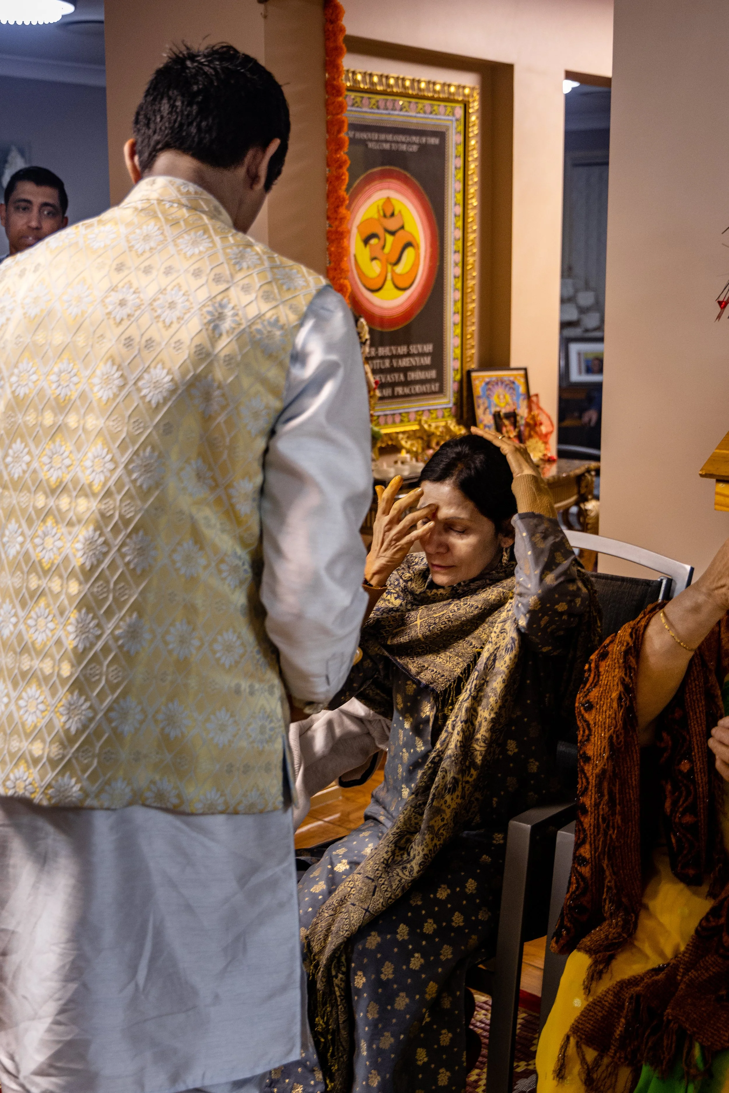 A woman sitting with her eyes closed and hands on her forehead, appearing distressed, while a man stands in front of her during a religious ceremony at a Hindu temple or prayer room decorated with religious pictures and symbols.