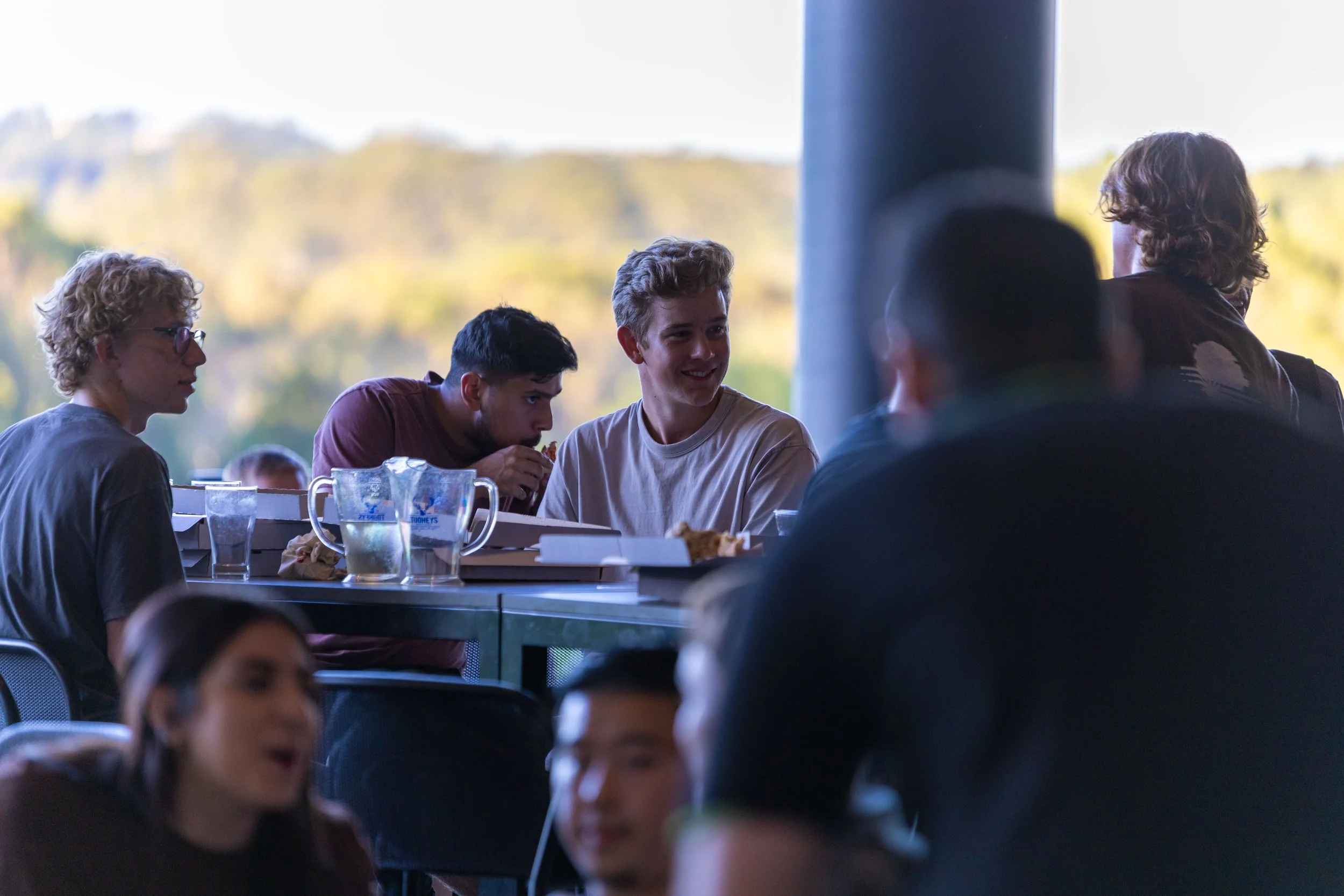 Group of young people sitting and eating together at an outdoor seating area, with a scenic autumn background.