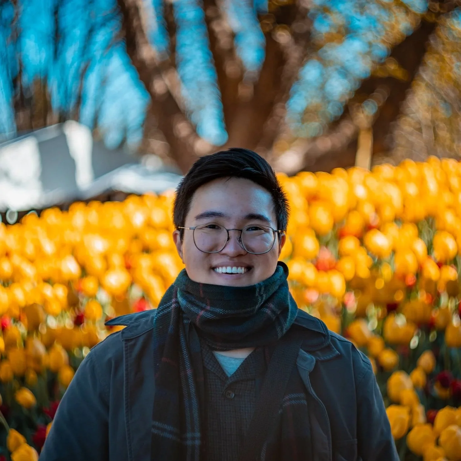 A smiling person with glasses and a scarf standing in front of a field of yellow flowers, with blurred trees in the background.