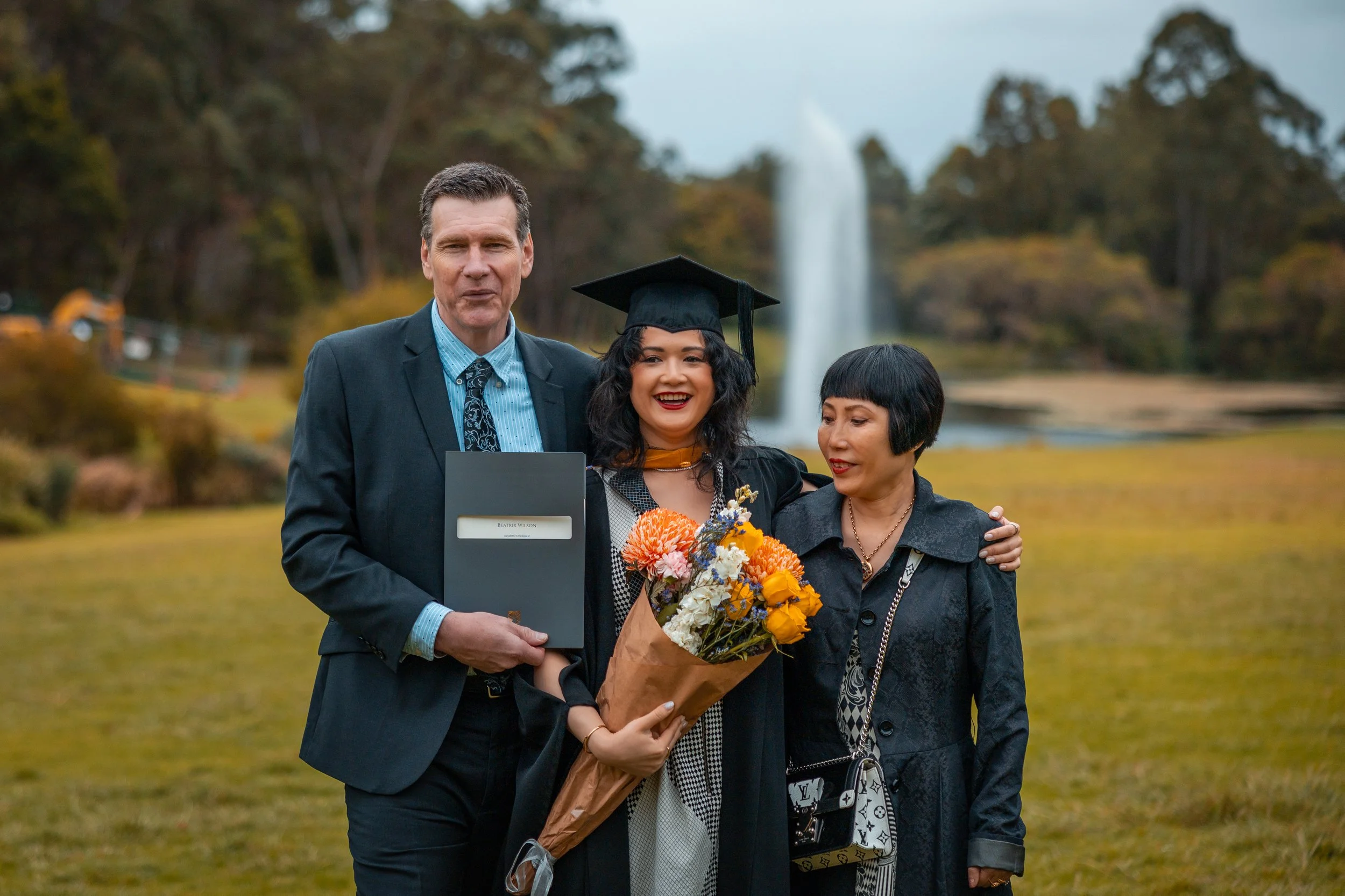 A young woman in graduation cap and gown holding a bouquet of flowers, standing with a man and a woman outdoors near a fountain and trees.