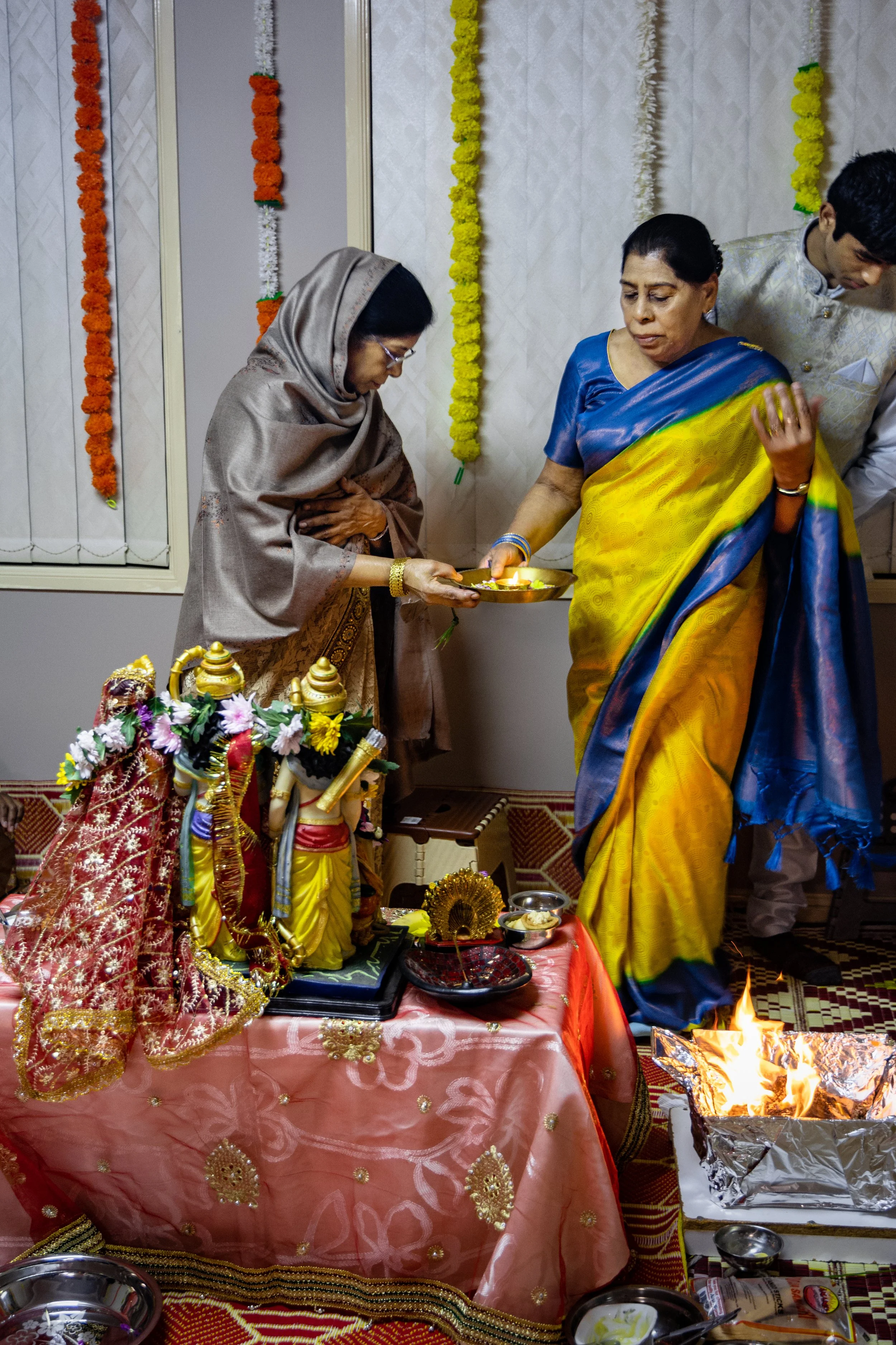 A traditional Indian religious ceremony with women in sarees performing rituals, prayer statues, and offerings on a decorated table.