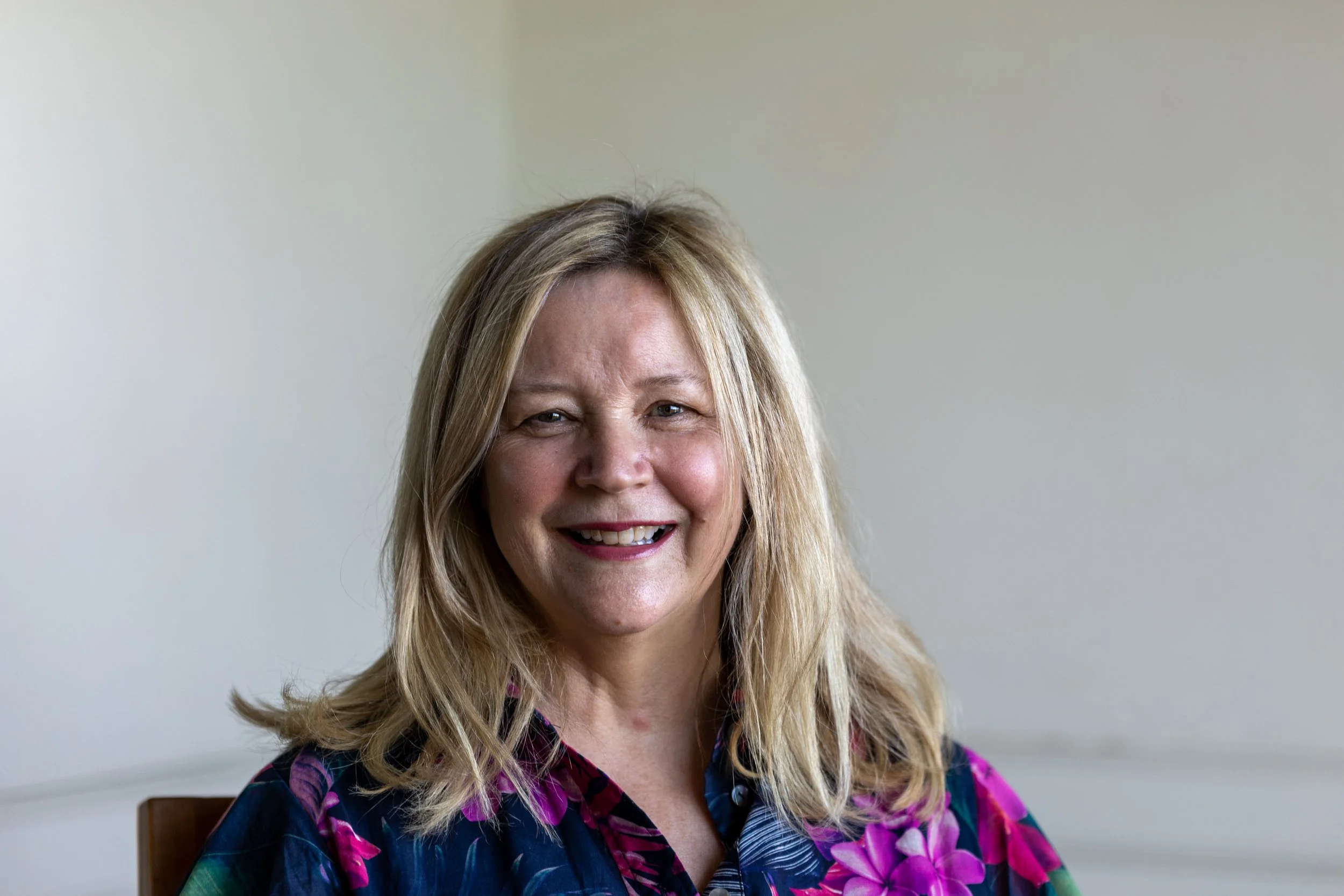 A smiling middle-aged woman with blonde hair wearing a colorful floral shirt, sitting against a plain light-colored wall.