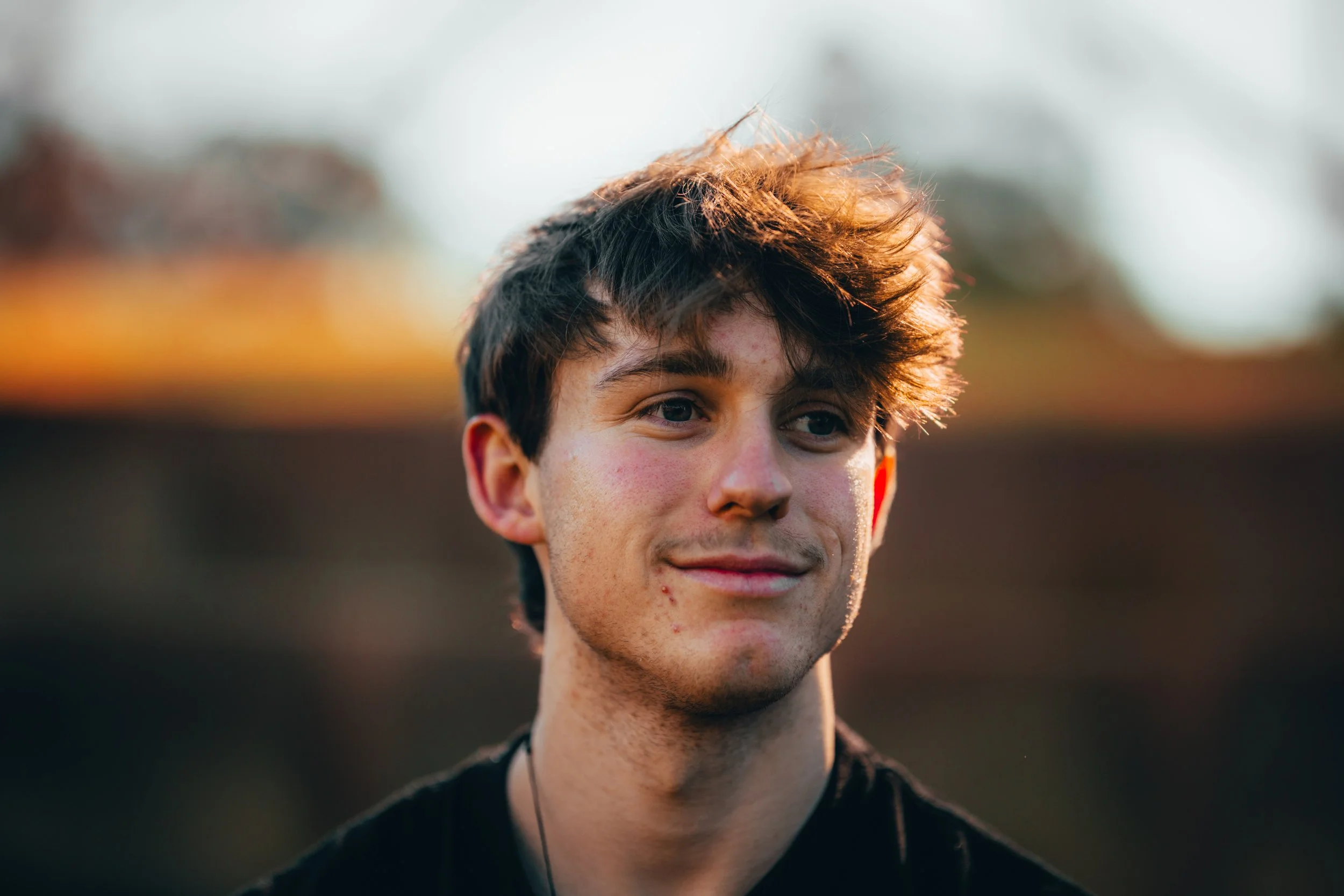 Close-up of a young man with tousled brown hair, fair skin, and a slight smile, outdoors during at sunset or late afternoon.