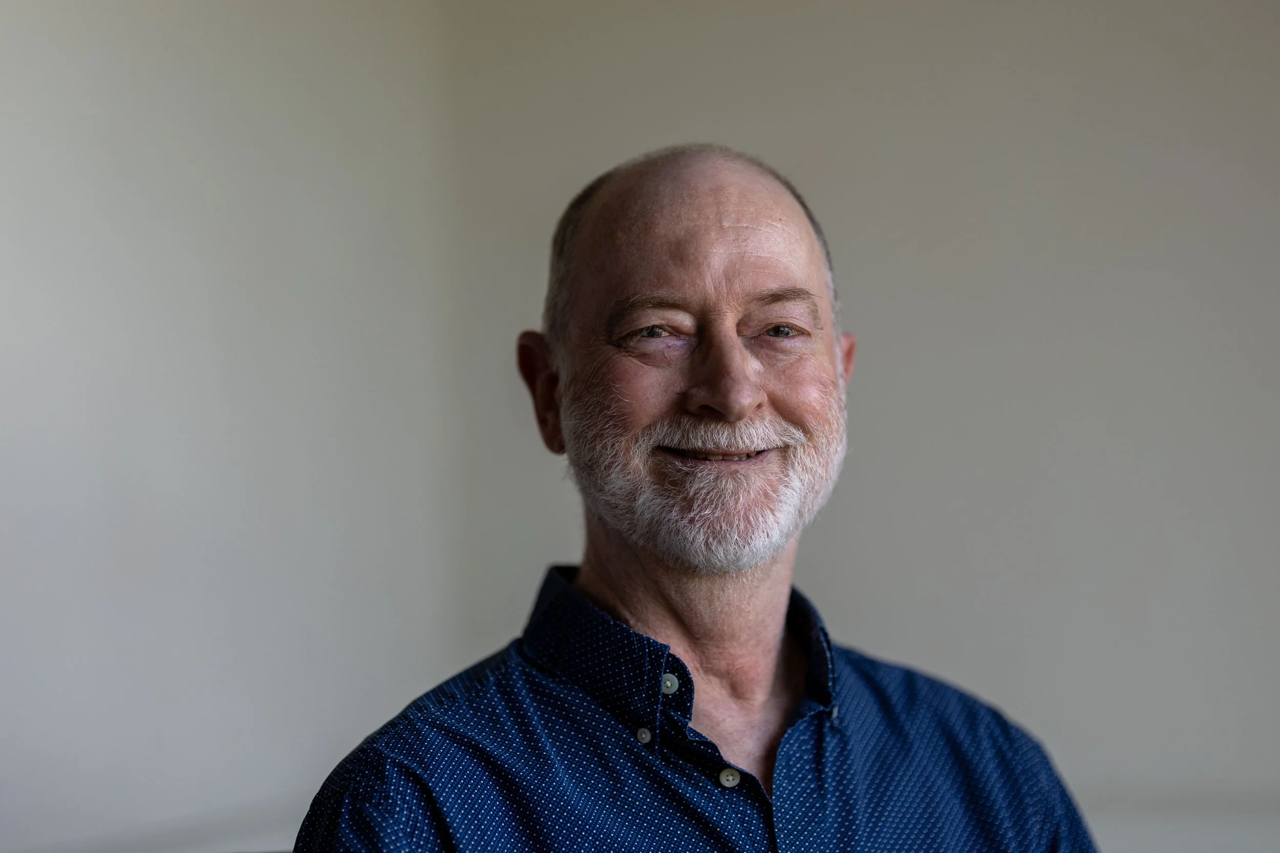 Portrait of an older man with a beard and mustache, smiling, wearing a dark blue shirt, against a plain background.
